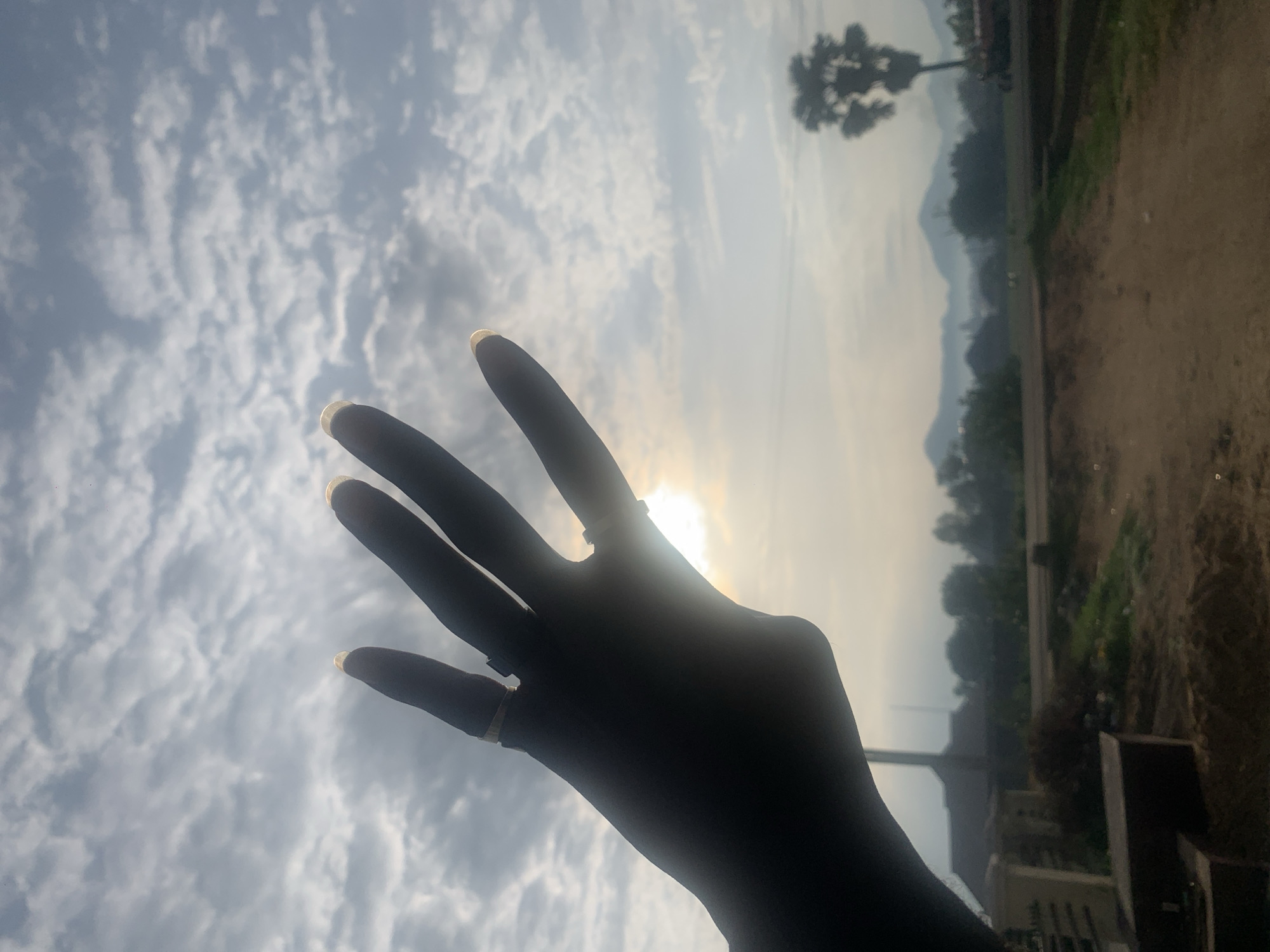A silhouette of a person's left hand is held up against a cloudy sky, with the sun partially visible behind the hand. The hand has two rings on it, one on the ring finger and another on the middle finger. The sky is filled with scattered, wispy clouds. In the background, there are faint outlines of hills or mountains, a lone palm tree, and some buildings that suggest a rural or semi-rural environment. The ground in front of the buildings appears to be a dirt path or road. The overall lighting suggests either sunrise or sunset. The location is Howayi, Nigeria.