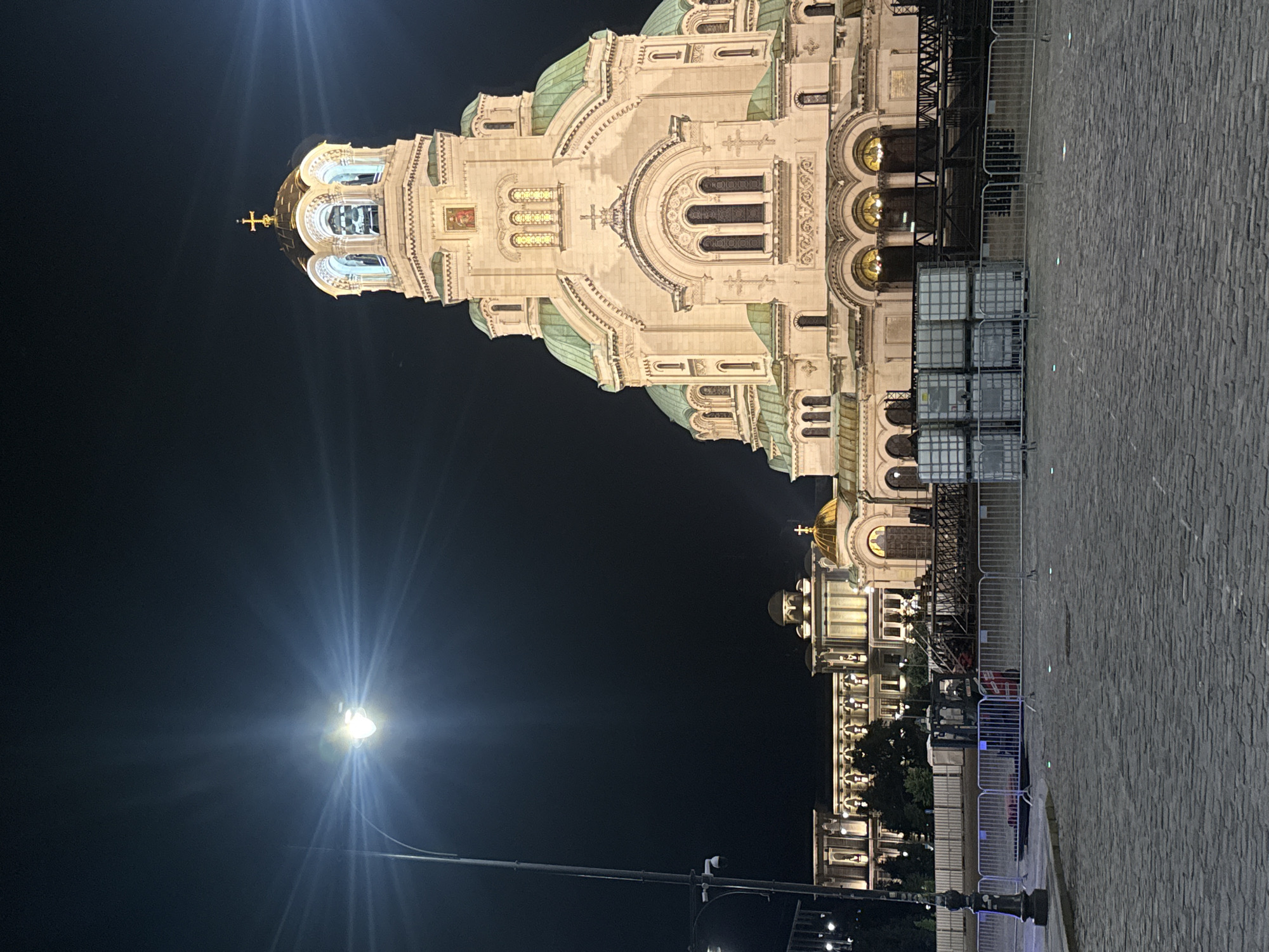 The image, taken at night in Sofia, Bulgaria, features the majestic Saint Alexander Nevsky Cathedral prominently displayed against a dark sky. The cathedral is brilliantly illuminated by artificial lights, highlighting its intricate architectural details.
The grand edifice showcases multiple domes, some with a distinctive greenish hue, characteristic of oxidized copper, topped with golden Orthodox crosses. The facade is adorned with ornate arches, columns, and decorative elements, including what appears to be an icon or mosaic embedded in an upper section of the building, as hinted by the crops.
In the foreground, a wide paved area, possibly cobblestones or large pavers, stretches towards the right. A bright streetlight on the left casts a strong starburst effect across the dark sky. In the mid-ground, a series of white metal barriers or fences are visible, along with several stacked, large white rectangular containers, possibly industrial bulk containers (IBC totes) or temporary structures. Dark scaffolding or support structures are also present near the base of the cathedral, suggesting ongoing work or preparations. A few trees are partially visible behind the barriers, blending into the dark background.
No people are visible in the scene. The overall impression is one of grandeur and historical significance, with the illuminated cathedral serving as the striking centerpiece against the night sky, possibly hinting at an event or maintenance in the area due to the barriers and containers. No legible text is visible in the image.