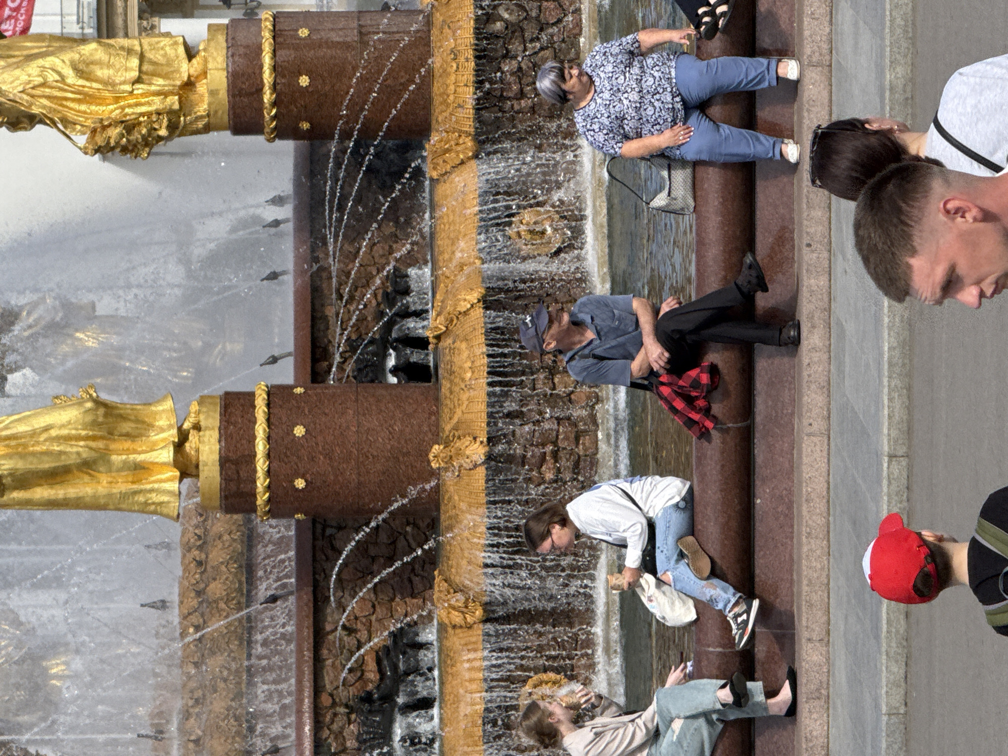 A group of people are gathered near a large, decorative fountain in Moscow, Russia. The fountain features ornate gold accents and cascades of water, with a rough stone texture forming the backdrop for the water flow. The scene is outdoors, likely during daylight hours given the brightness.
Several individuals are visible in various states of repose and interaction. In the upper portion of the image, a woman with short gray hair wearing a blue and white patterned shirt and blue pants sits with her legs crossed. Next to her, a person in a grey shirt and dark pants is partially visible. Further down, a man in a blue shirt and dark pants, wearing a baseball cap, sits near the fountain. A woman in a white shirt and distressed blue jeans is also present, looking down towards something in her hands. In the lower part of the frame, a person in a light-colored jacket and light blue pants is seated on the ground, looking at their phone. Another person, wearing a red hat, is partially visible on the right side of the image. The overall mood appears relaxed, with people enjoying the fountain area.
Notable details include the detailed gold embellishments on the fountain's structure, the way the water flows and glistens, and the casual attire of the people. The presence of a fountain of this scale suggests a public park or plaza. There is no visible text in the provided image.