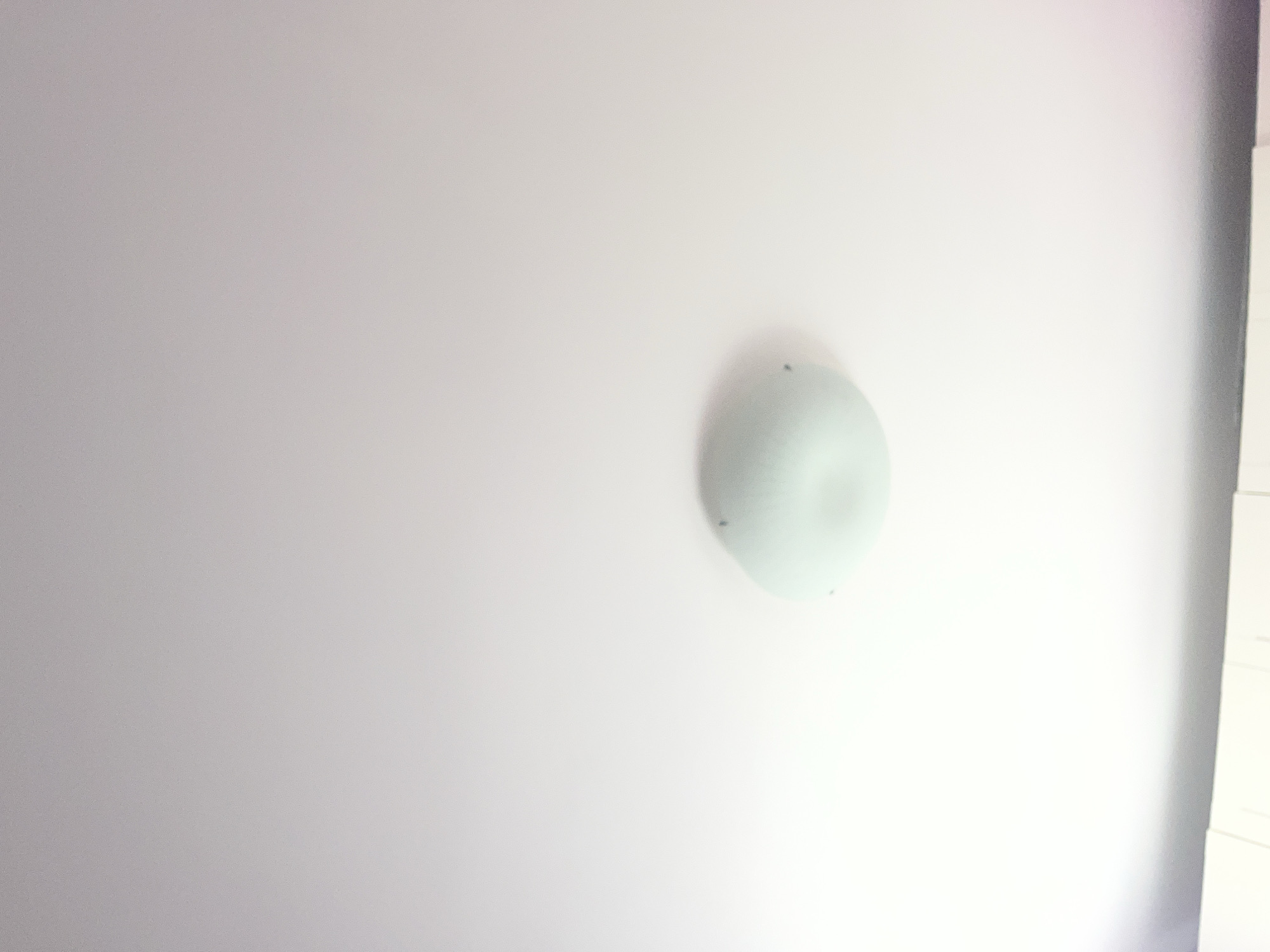 The image shows a white ceiling with a round, semi-transparent light fixture affixed to it. The wall is white and flat, suggesting an indoor environment. The upper right corner shows a partially visible structure, possibly a wall, with a linear pattern, suggesting panels or sections. The scene appears to be indoors and artificially lit. The location of Pokrovnik, Bulgaria is irrelevant as there are no landmarks or attributes to confirm the location in the image.