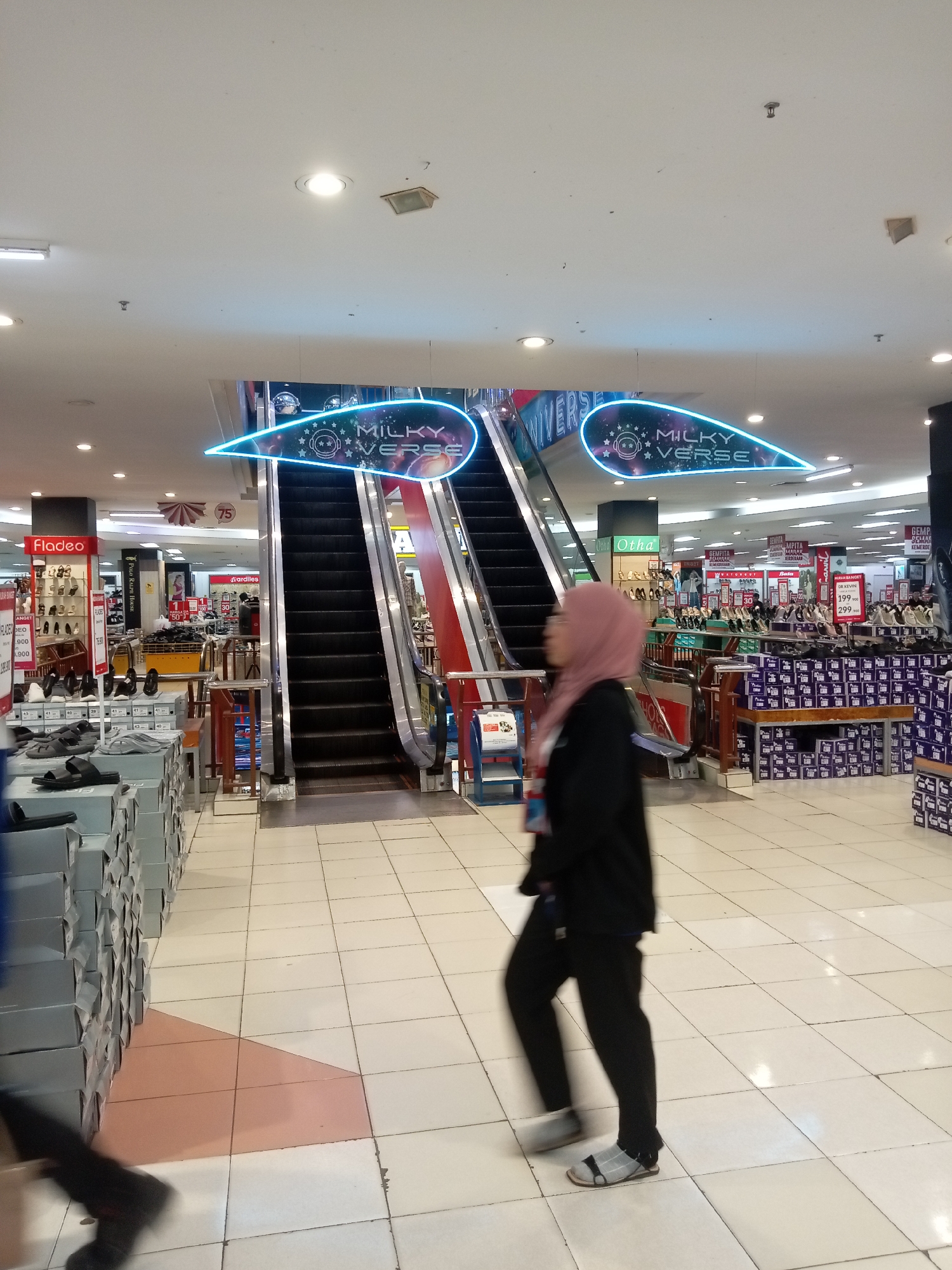 The image depicts the interior of a department store, likely in Batam, Indonesia, with a focus on shoes. In the foreground, a person wearing a hijab and a black jacket is walking away from the camera, their movement blurred. The floor is tiled with white squares and some lighter brown tiles arranged in a curved pattern. To the left, stacks of shoe boxes are visible, some open, displaying various sandals and shoes. A sign for "Fladeo" is prominent on a display shelf.
In the mid-ground, two escalators ascend, with a neon blue sign above them reading "MILKY VERSE." The escalators are positioned in front of a wide open space within the store, revealing more aisles and displays of footwear. Signs for brands like "Otha," "Bata," and "Ardiles" are visible, along with price tags and promotional materials indicating sales and discounts, such as "DISCOUNT 50%" and prices like "199.900" and "299.900".
The background shows a typical retail environment with bright overhead lighting, more shelves stocked with merchandise, and other shoppers in the distance. The overall impression is that of a busy shoe section within a large retail establishment.