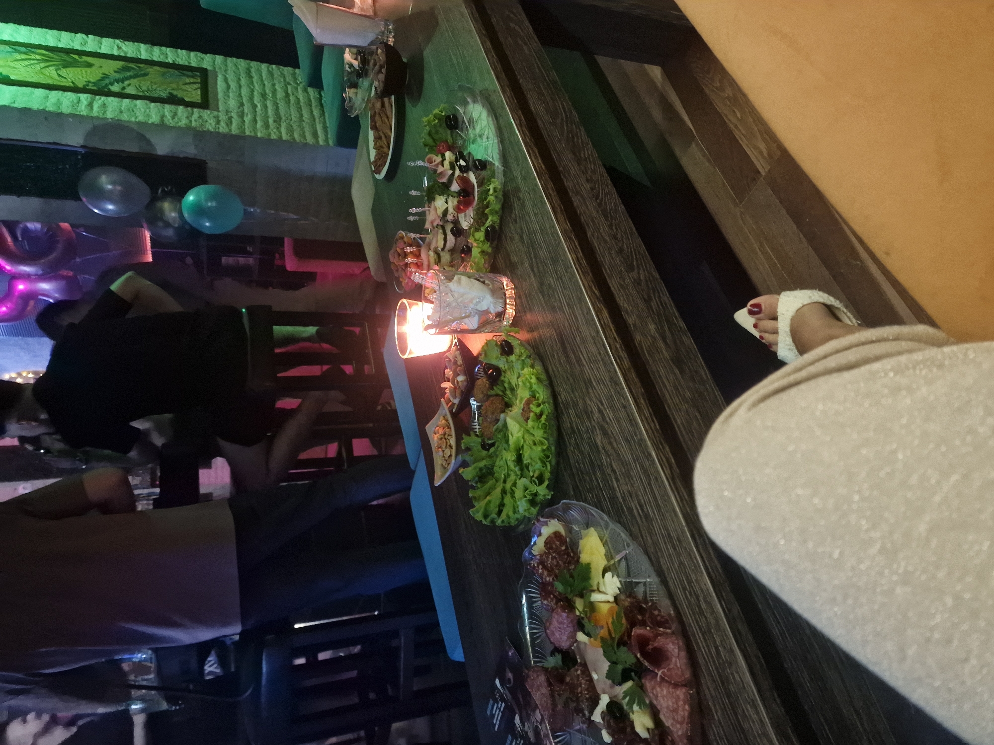 The image depicts a dimly lit indoor scene, likely a party or gathering, with a focus on a table laden with food. Several people are present, though their faces are not clearly visible. One individual, seen from the waist down, is wearing a light-colored robe and white fuzzy slippers with red nail polish.
The table is covered with various food platters. One platter contains what appears to be a charcuterie board with an assortment of sliced meats, cheeses, and garnishes of parsley and olives. Another platter features skewers of meat and what looks like a side of lettuce. There are also small dishes of nuts and possibly dips. A lit candle in a glass holder provides a warm glow.
In the background, there are balloons, one teal and another silver, suggesting a celebratory occasion. The walls appear to be textured, with some artwork or decorative panels visible. The overall atmosphere is casual and social. The lighting is low, with colorful accents from some unseen light sources, contributing to a festive and somewhat intimate mood. There is no visible text in the image.
