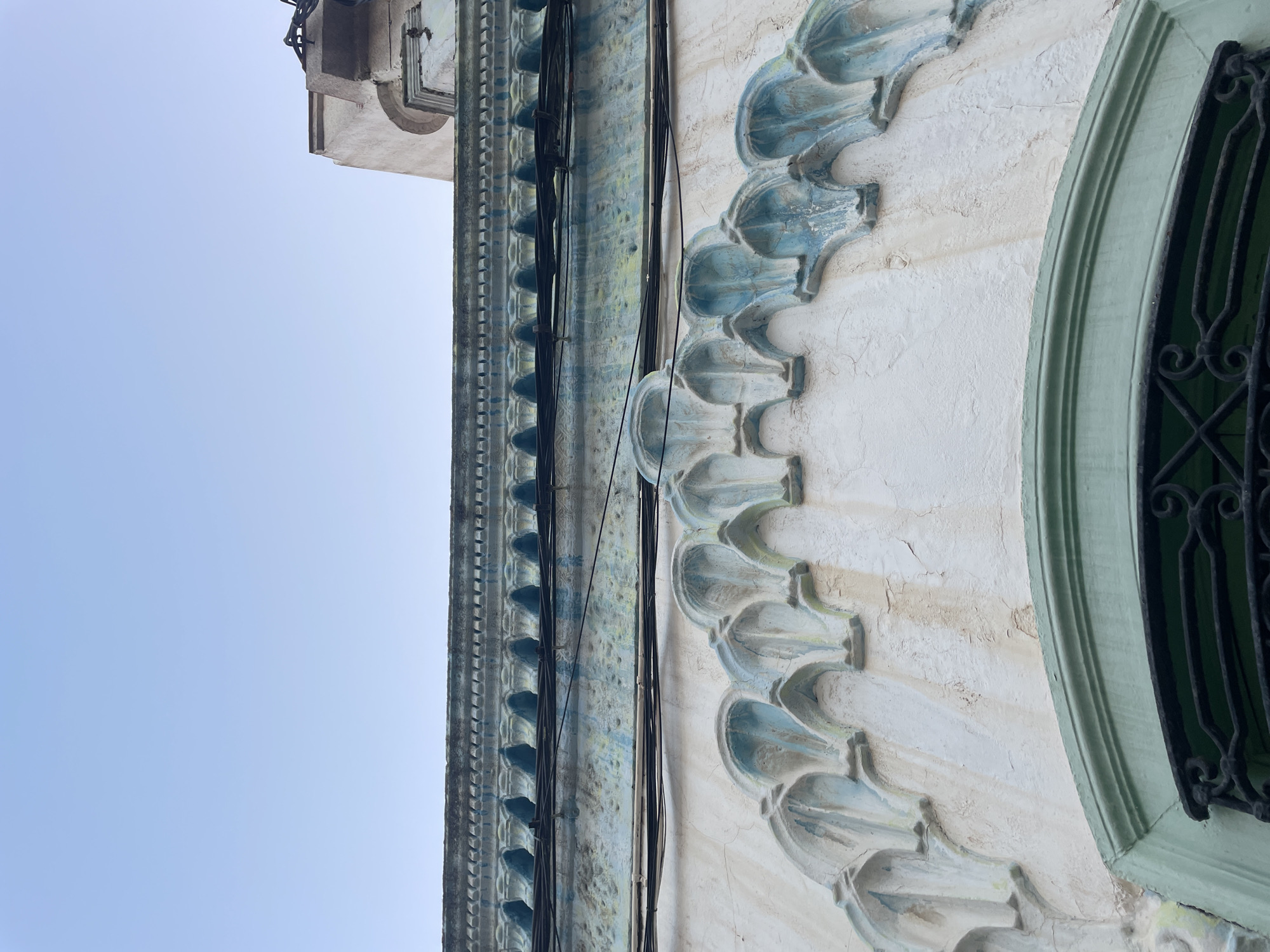 This is a low-angle, exterior shot of a building's facade in La Línea de la Concepción, Spain. The sky is clear and a pale blue, taking up the top half of the image. The building features architectural details with a distressed, aged appearance.
The upper part of the facade has a band of decorative molding with a textured, almost serrated edge, painted in a faded teal color. Below this, a horizontal line of dark electrical wires runs across the wall. Further down, a series of arch-like decorative elements are prominently featured. These elements are painted in a light cream color, with accents of pale teal and hints of yellowing and water stains, suggesting age and weathering. The texture of the wall is rough and uneven.
At the bottom of the image, the curved top of a window or doorway is visible, framed by a green-painted border. Below this frame, a dark, ornate wrought-iron grille is present, partially obscuring what lies beyond. The overall impression is of an old building with intricate, historical architectural features that show signs of wear and tear.