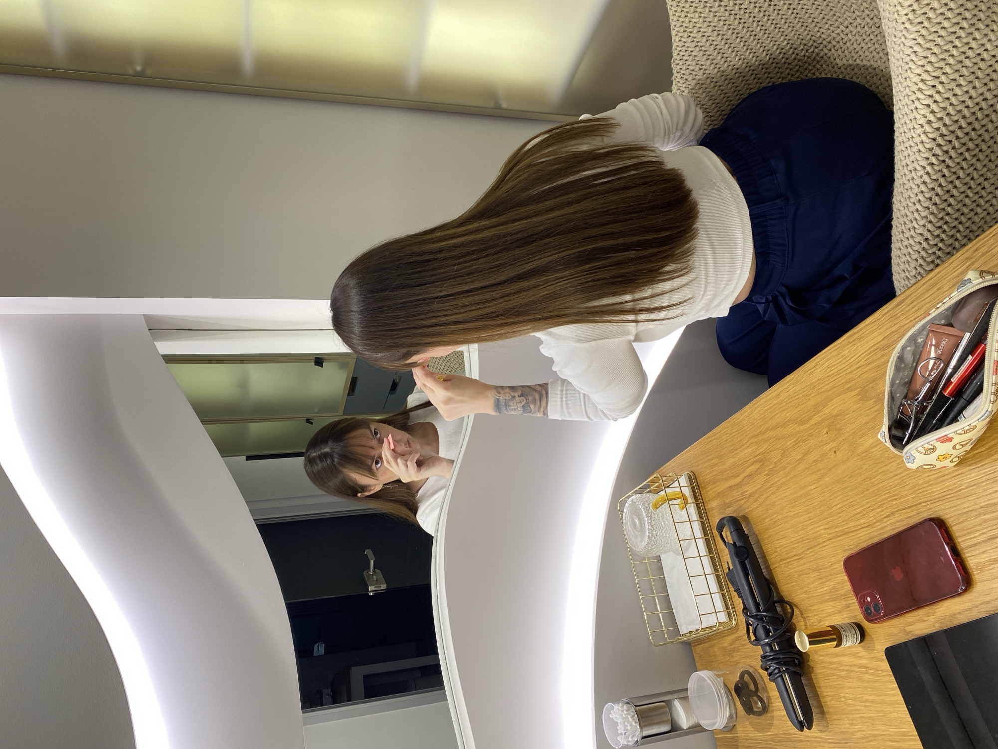 A woman with long brown hair, wearing a white top and blue pants, sits in front of a vanity mirror. Her reflection shows her smiling and holding something up to her face. The vanity has a wooden countertop with a makeup bag, a gold wire basket holding a glass and some tissues, a black curling iron, a small bottle, a burgundy cell phone, and a black laptop. The environment is a room with gray walls and a white curved light fixture around the mirror. There is no information in the image to confirm the location is in Votkinsk, Russia.