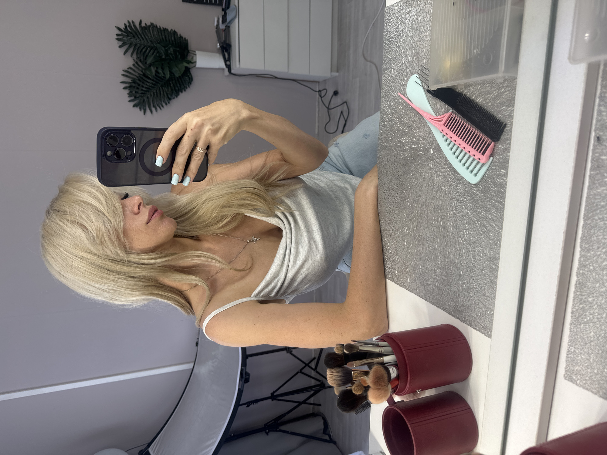 This media file shows a woman with blonde hair taking a mirror selfie. She is wearing a light gray tank top and light blue jeans. She is holding a black smartphone in her right hand, which is also adorned with a ring. Her fingernails are painted a light blue color. Her left arm is resting on a surface, and her body is angled towards the camera.
In the background, a white cabinet with gray wooden paneling is visible. To the left of the woman, there are two artificial fern leaves in a white vase. To the right of the woman, on a textured silver surface, there are two combs, one pink and one black. A red cylindrical container holding makeup brushes is also visible in the foreground. A softbox light is partially visible at the bottom left of the image. The overall setting appears to be an indoor space, possibly a dressing room or a vanity area. The lighting suggests it could be daytime, as there's natural-looking light. The woman's expression is neutral, and she appears to be focused on taking the picture. There is no visible text in the image.