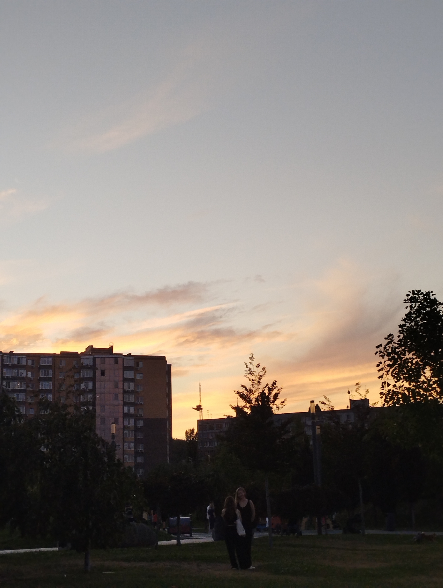 The image shows a sunset over an urban area. Two women are standing in a grassy park in the foreground. Behind them, a street with trees and lampposts is visible, and further in the background, a tall apartment building and a construction crane are silhouetted against the orange and pink sky. The overall lighting suggests it is dusk.