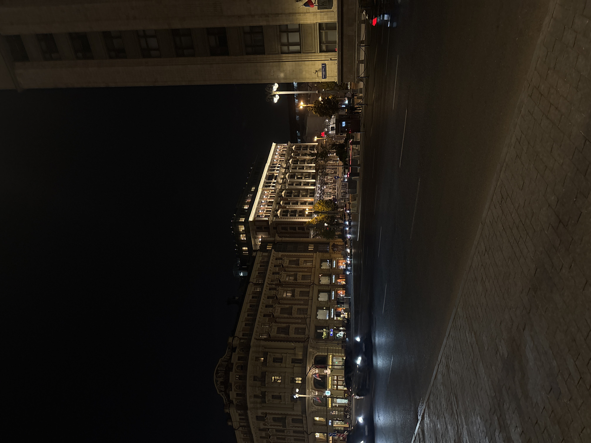 The image depicts a night scene on a city street in Moscow, Russia. A grand, illuminated building, likely a hotel or significant architectural landmark, dominates the left side of the frame. Its facade is adorned with numerous lit windows and decorative elements. Across the street, a tall, imposing building with a classical architectural style stands on the right, partially obscuring the dark night sky. Cars with their headlights on are driving on the wet asphalt road, suggesting recent rain or a damp surface. The sidewalk in the foreground is paved with brickwork. Streetlights cast pools of light, and a few trees are visible, adding a touch of nature to the urban landscape. The overall atmosphere is one of a bustling city at night, with the architecture and lighting creating a dramatic and sophisticated ambiance.