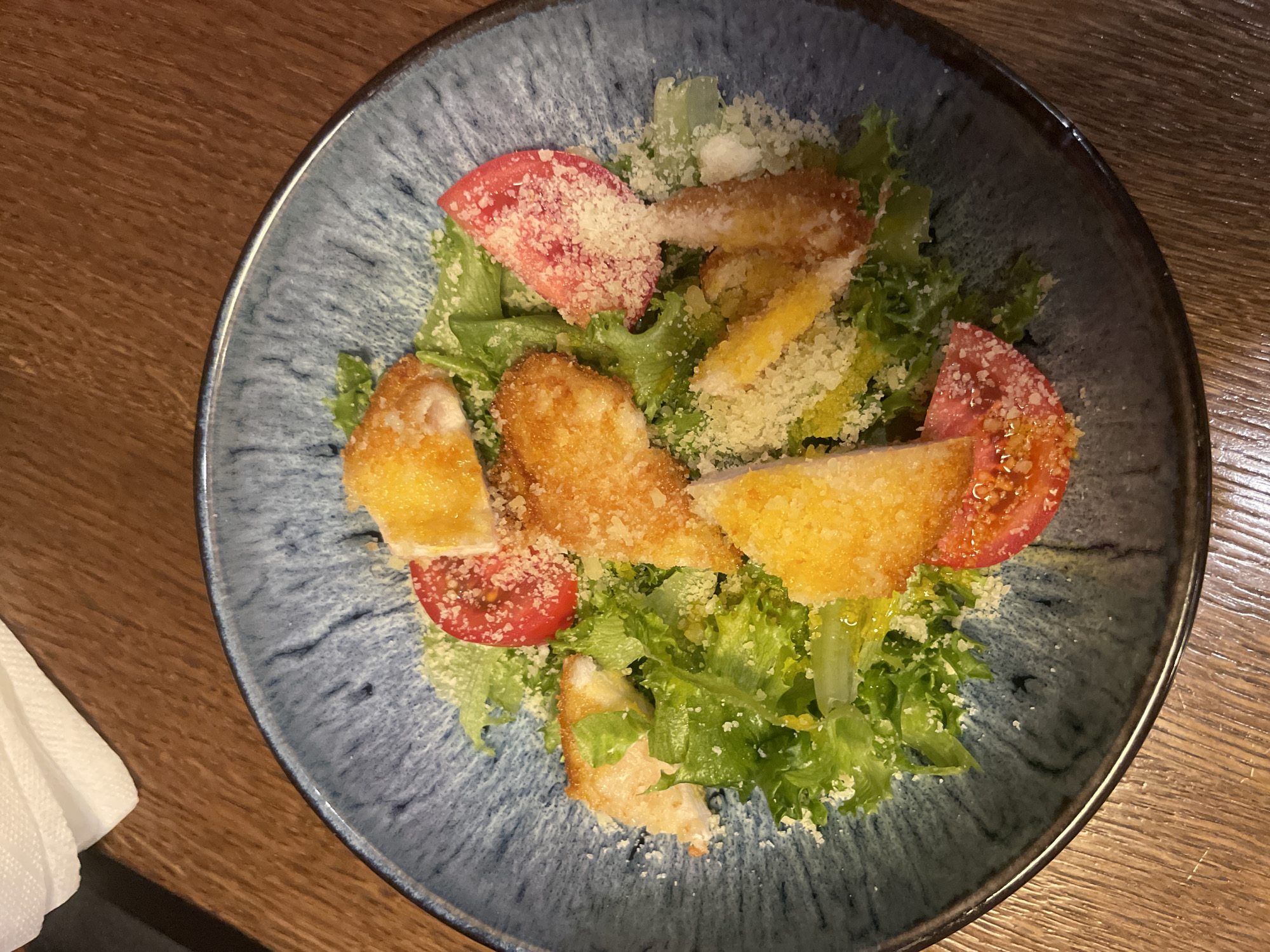 A top-down view shows a blue-grey ceramic plate containing a salad. The salad ingredients appear to be green lettuce, sliced red tomatoes, breaded chicken or fish, and grated cheese. A white napkin is partially visible at the bottom left of the image. The plate sits on a wooden table. The image appears to be taken in a restaurant setting. The specific location in Izhevsk, Russia, cannot be determined from the image.