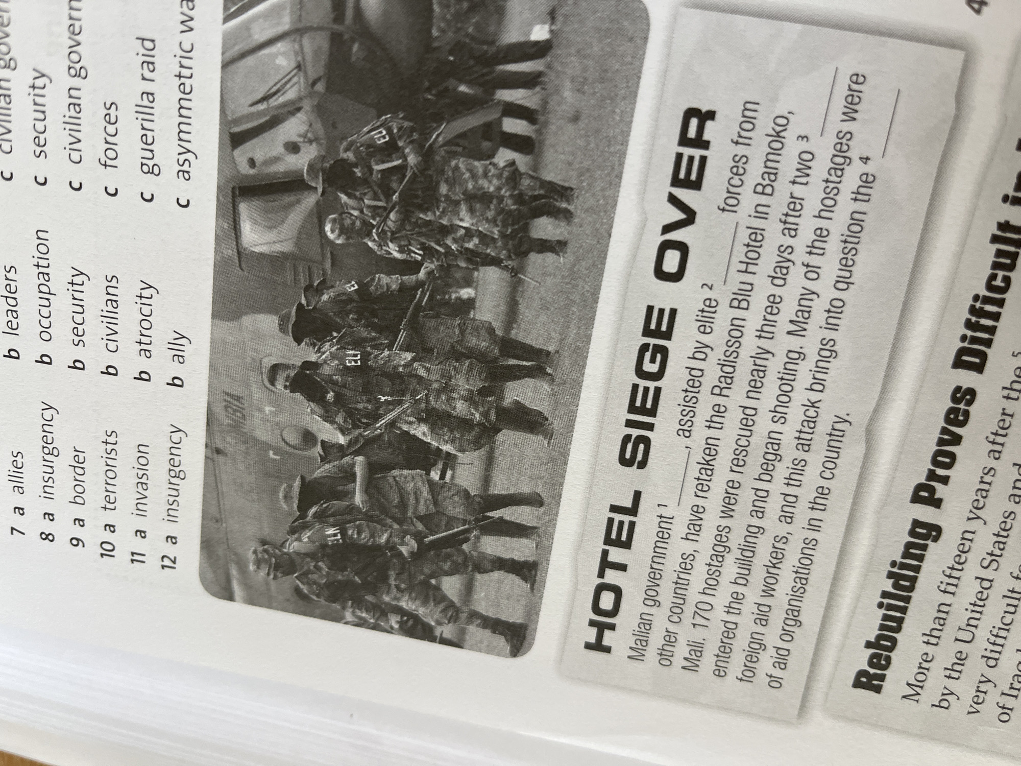 The image is a page from a textbook or workbook, featuring a news report about a hotel siege in Bamako, Mali. The title reads "HOTEL SIEGE OVER". The text describes how the Malian government, with assistance from elite forces and other countries, retook the Radisson Blu Hotel. Around 170 hostages were rescued nearly three days after two foreign aid workers entered the building and began shooting.
Beside the article is a black-and-white photograph depicting soldiers in camouflage uniforms, possibly involved in the operation. The bottom right corner of the workbook page has another news title: "Rebuilding Proves Difficult".
There is also a multiple-choice section with vocabulary related to conflict.
There is nothing in the image to indicate that the location is the city of Panagyurishte, Bulgaria.