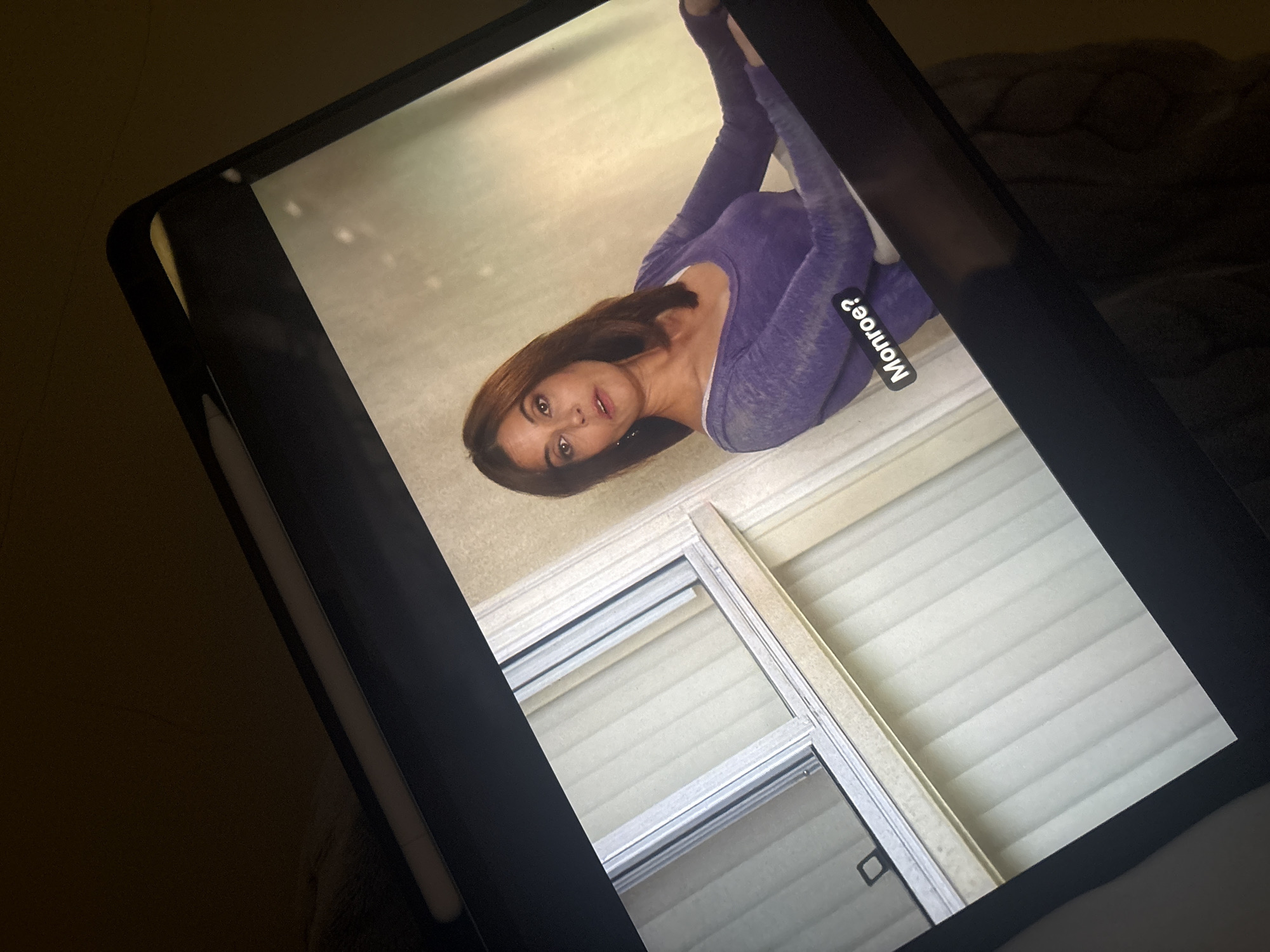A woman in purple athletic wear is lying on the floor in an Unknown location, as viewed on a tablet. The background includes blinds, and a text overlay says "Monroe?". A stylus is beside the tablet.