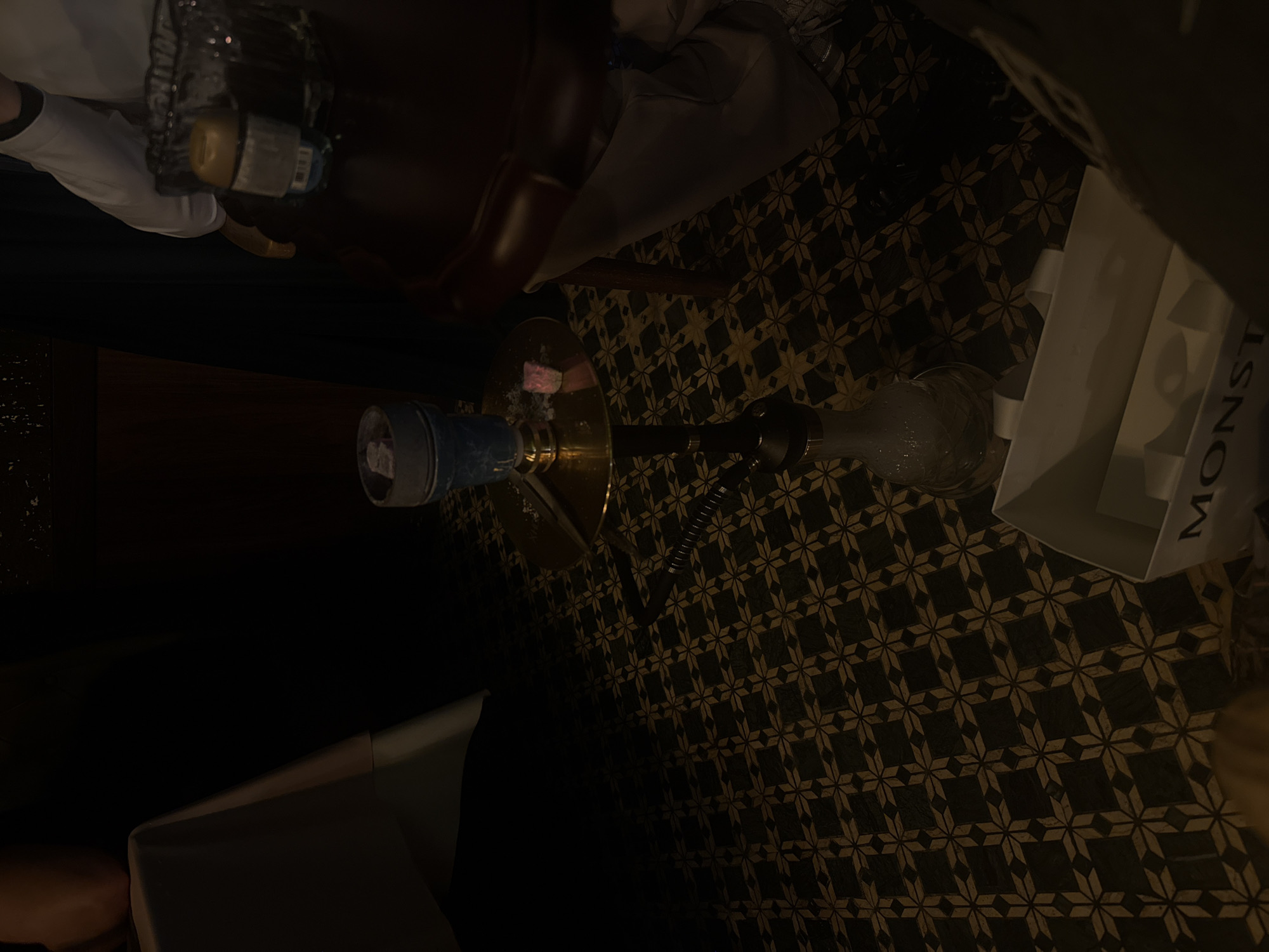 The image depicts a hookah setup on a dark, geometrically patterned surface, possibly a floor or tabletop. A blue and silver cylindrical object (likely the hookah base or filter) is connected via a hose to a glass water chamber. Above the hookah is a dark brown object, possibly a table or chair. In the upper right corner, a white box with lettering (partially obscured, but appears to say "MONST") is visible. A person's arm is visible in the upper left. The photo appears to be taken indoors under dim lighting. There is no explicit indication of location in Haizhu District, China.