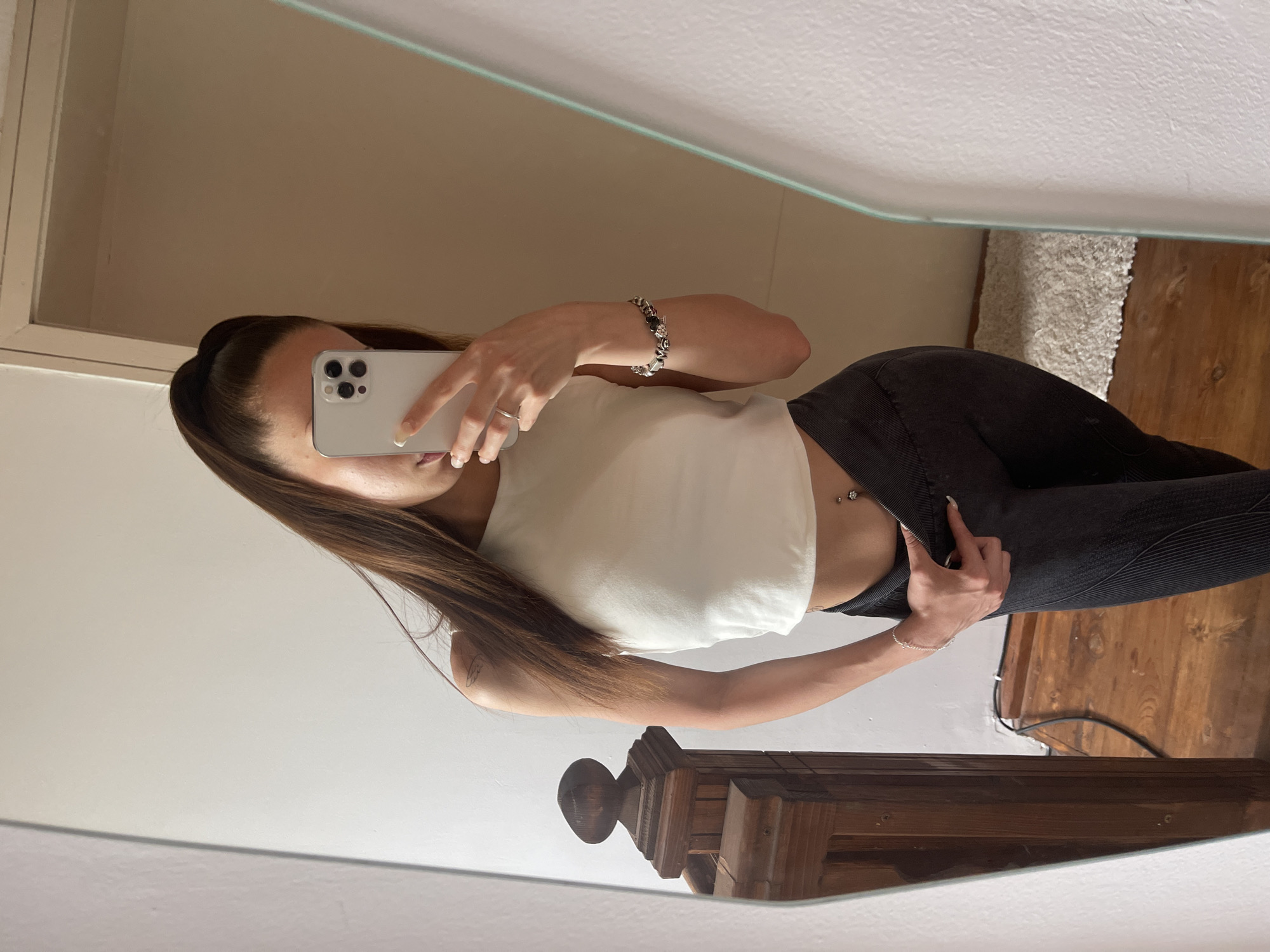 In an indoor setting, a young woman with brown hair pulled into a high ponytail is reflected in a mirror, taking a selfie with her phone. She is wearing a white crop top and dark leggings. Her right hand is visible, holding the phone, and she wears a ring and a bracelet. A navel piercing is visible. To her left, a wooden railing is reflected in the mirror. The background consists of a white wall and a wooden floor. The location is described as Unknown, Unknown.