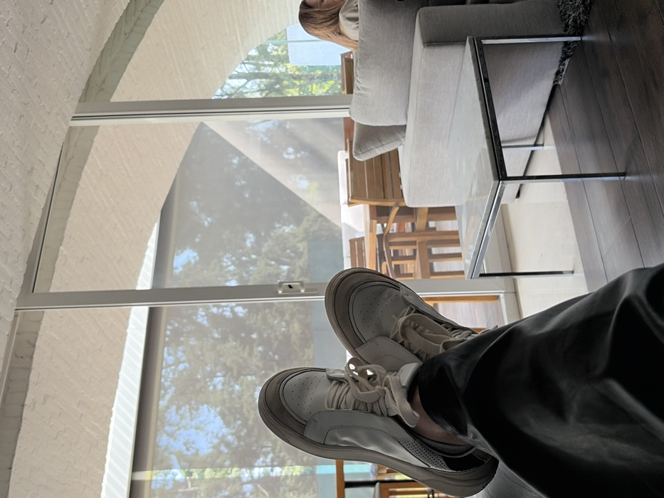 The image captures an indoor scene, likely a modern living room or lounge area, with a person's legs and feet prominently in the foreground, indicating a first-person perspective of someone relaxing. The image is oriented with the floor on the right side and the ceiling/archway on the left, suggesting it was originally taken with the camera rotated.
In the immediate foreground, a person's legs, clad in dark pants (possibly black or dark grey), are visible, extending towards the viewer. They are wearing a pair of white sneakers with visible laces and some perforations on the toe box. One shoe also shows a reflective element on the heel area.
The background reveals a spacious room bathed in natural light. To the left, a white brick wall or archway is visible, suggesting architectural detail. Large windows occupy a significant portion of the scene, fitted with translucent roller shades that diffuse the sunlight. Through these shades, the outlines of trees and a bright blue sky can be seen, indicating it is daytime. Outside the window, a glimpse of outdoor wooden furniture, including a table and chairs, is discernible.
To the right of the image, a light grey fabric sofa with cushions is partly visible. Adjacent to the sofa is a modern coffee table with a glass top and a thin, dark metal frame. The floor in this area is a dark hardwood. In the upper right quadrant, partially obscured by the sofa, a person (likely a woman with light-colored hair) is visible, looking away from the camera, adding a subtle human element to the scene.
The overall impression is one of leisure and comfort in a bright, contemporary setting, with a connection to the outdoors through the large windows. No text is visible in the image.