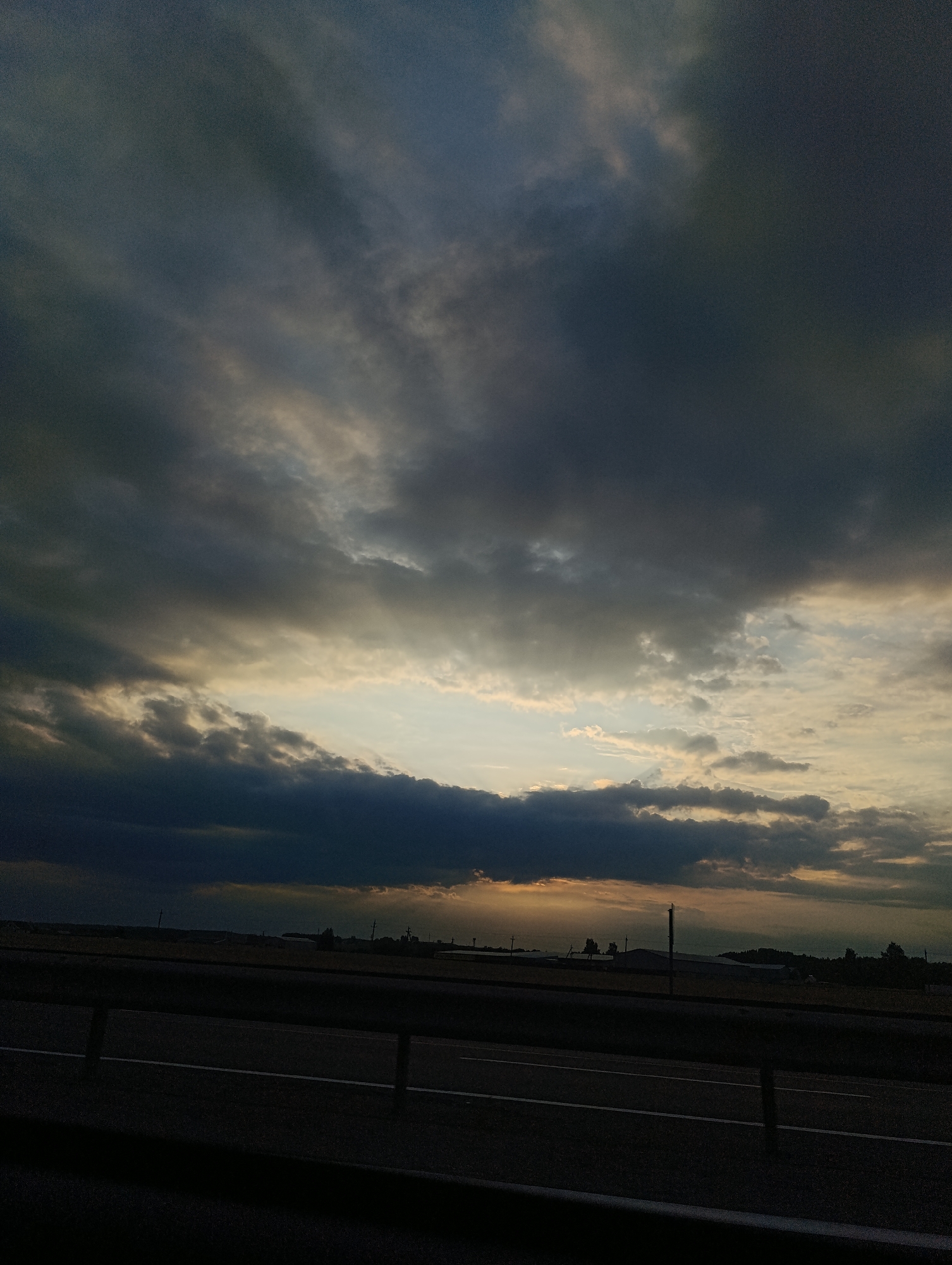 The image shows a view of a rural landscape at dusk, as seen from the side of a road. The sky is filled with dramatic, dark clouds, with a sliver of golden light breaking through near the horizon. This light illuminates the tops of some of the clouds and casts a warm glow on the land in the distance. The foreground is dominated by a dark, blurred expanse which appears to be the road and a guardrail, with white lines indicating lanes. In the background, silhouetted against the sky, are a few low-lying buildings and some scattered trees, suggesting a small settlement or agricultural area. The overall impression is one of impending weather or the transition from day to night in a remote setting.