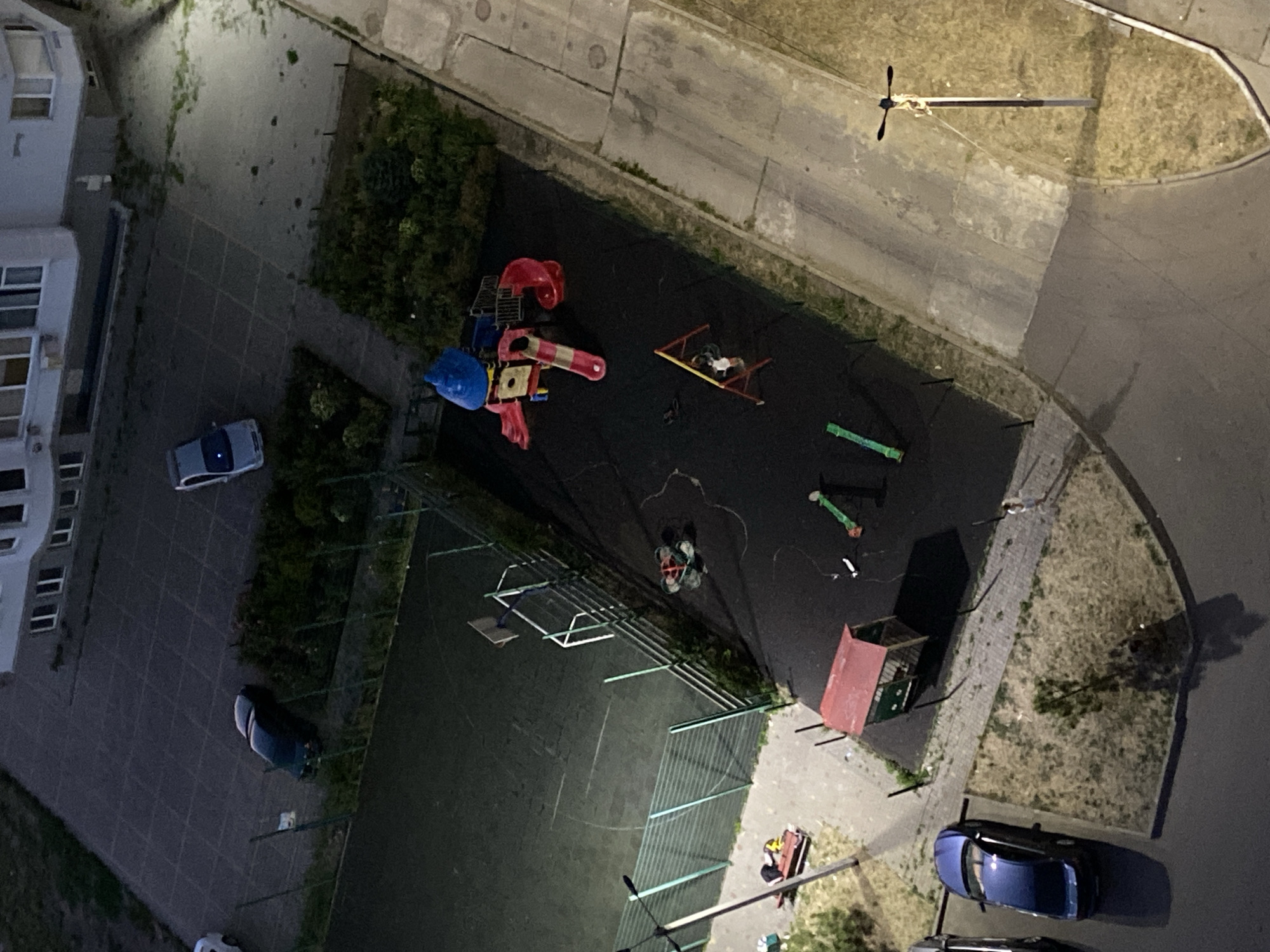 The image captures an overhead view of an urban outdoor scene, likely a residential courtyard in Chornomorsk, Ukraine, during twilight or low light conditions, as evidenced by the artificial lighting and shadows.
The scene is divided into several distinct areas. On the left, a dark green car is parked on a paved surface adjacent to a fenced-in sports court, possibly a basketball or football field, which is largely dark. A portion of a fence surrounds this court. Further left, a dark blue car is parked near a sidewalk.
Dominating the central area is a children's playground. It features a multi-colored play structure with slides, a yellow platform, and a blue canopy. A red, A-frame swing set is also present, with one figure, appearing to be a child, on it. Nearby, a small red-roofed structure, possibly a playhouse or storage unit, is situated. Scattered on the playground surface are several pieces of equipment, including what appear to be balance beams.
To the right of the playground is a paved pathway or road, bordered by a grassy area with a street lamp. Further to the right, more paved surfaces and grassy areas are visible, along with part of a building in the background. The building appears to be a multi-story residential complex with multiple windows.
There are indications of human presence: a figure on the swing, and another figure near the red-roofed structure, possibly interacting with it. A person is also visible walking on the paved area to the far right. Several cars are parked in various locations. The overall impression is a quiet evening in a typical urban setting.