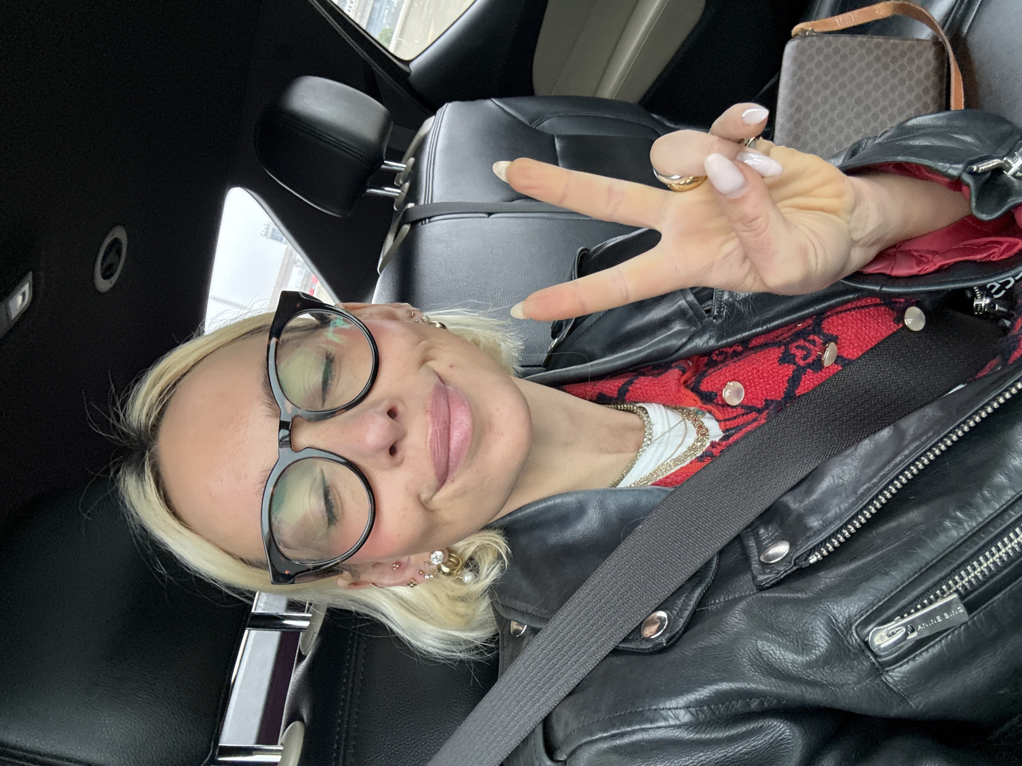 A blonde-haired woman wearing glasses and a black leather jacket with a red shirt underneath is making a peace sign while sitting in a car. The woman is wearing a seatbelt. A purse is visible in the background. The location is in a city, presumably New York City, United States of America.