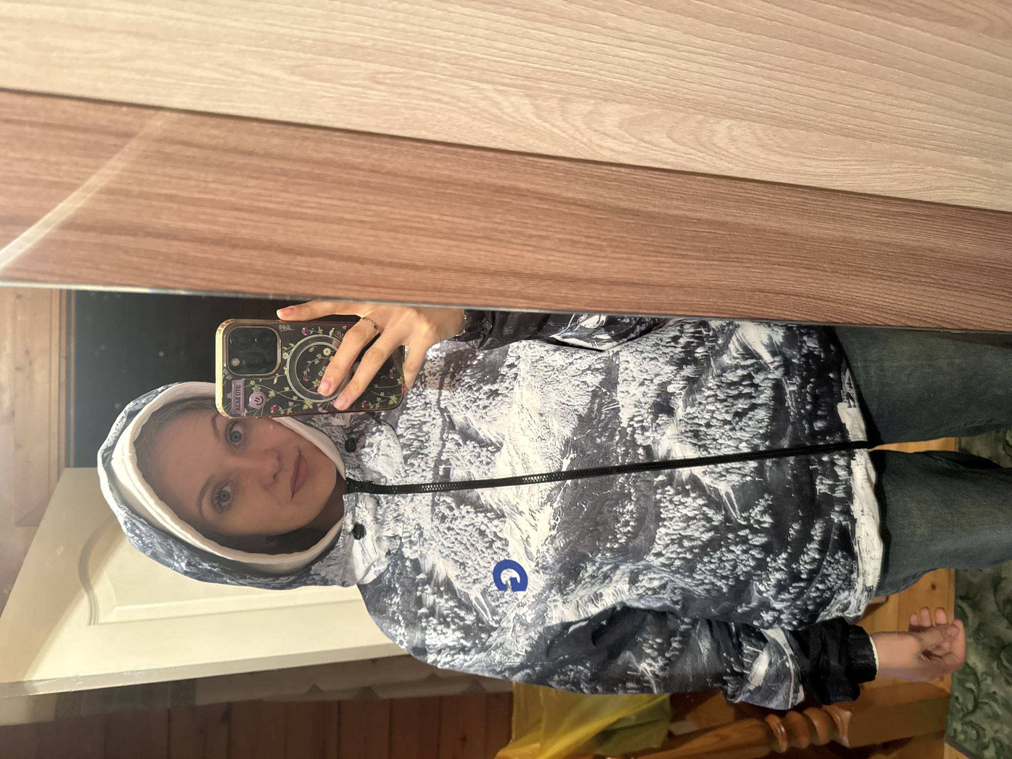 A woman takes a mirror selfie indoors. She is wearing a hooded jacket with a black and white mountain pattern and a blue "G" logo on the front. She is holding a phone with a floral case, and her face is visible in the reflection. The background includes a wooden surface (likely a wall or furniture) and a partially visible door. The woman's clothing includes dark jeans. The location cannot be confirmed with the information provided.