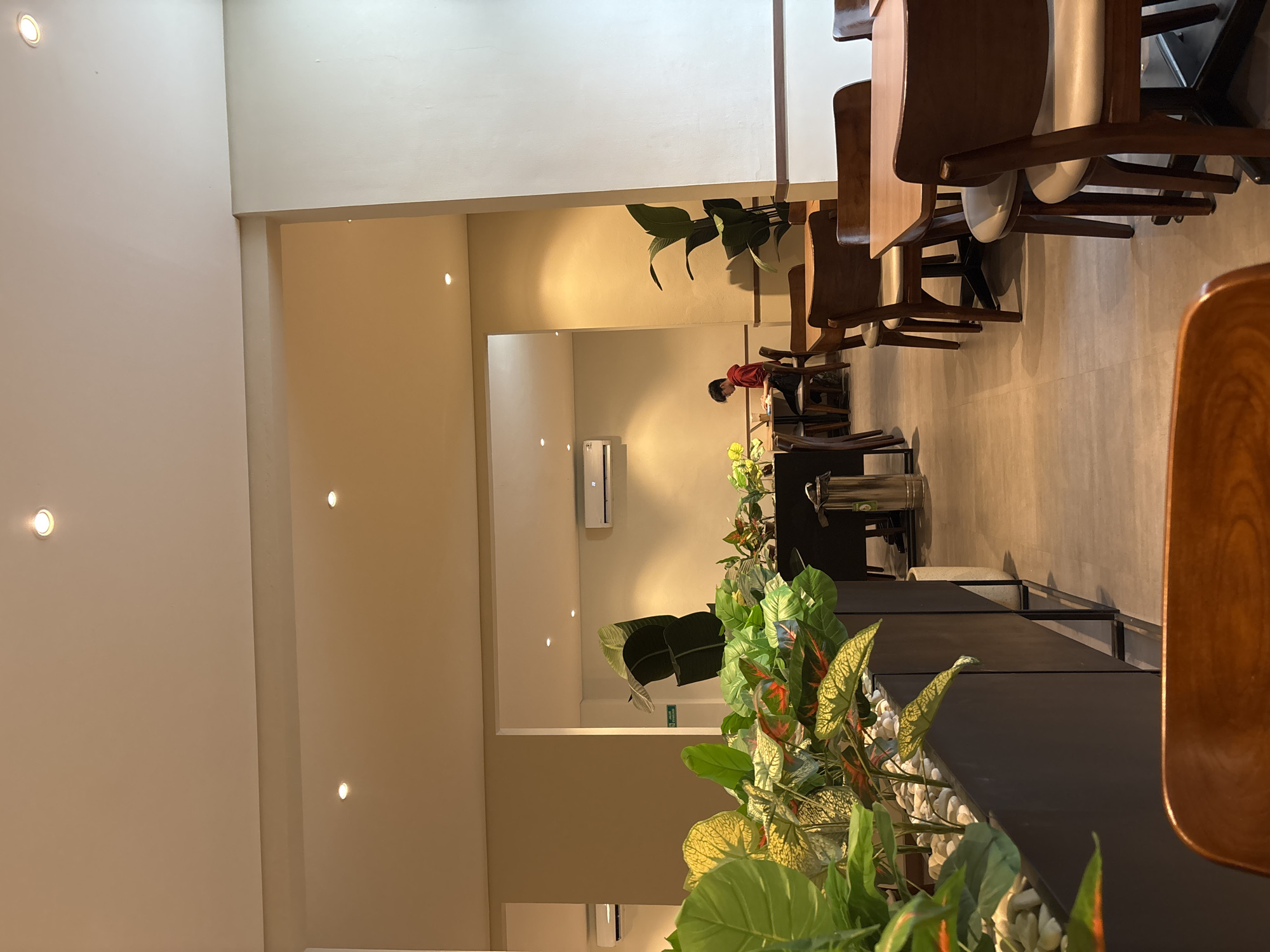 This is an indoor image of a dining area with tables and chairs, likely a cafe or restaurant, in West Jakarta, Indonesia. The scene is illuminated by ceiling lights and natural light filtering in from an open doorway.
In the foreground, lush green plants in a dark rectangular planter are visible. The plants have broad leaves with yellow and red variegation. The planter is filled with white stones.
Further into the scene, several tables and chairs are arranged. The tables are square or rectangular with light brown wooden tops and dark metal bases. The chairs are made of dark wood with light-colored upholstered seats.
In the middle ground, a young person wearing a red hooded sweatshirt is seated at a table, appearing to be working on a laptop or tablet. The person is facing away from the camera.
In the background, through an opening in the wall, another section of the establishment is visible with more tables and chairs. There are also air conditioning units mounted on the walls in both sections. Large plants with dark green leaves are placed in corners of the space.
The floor is tiled in a neutral grey color, and the walls are painted in light, neutral tones. The overall impression is a clean, modern, and well-lit interior space designed for dining or working.