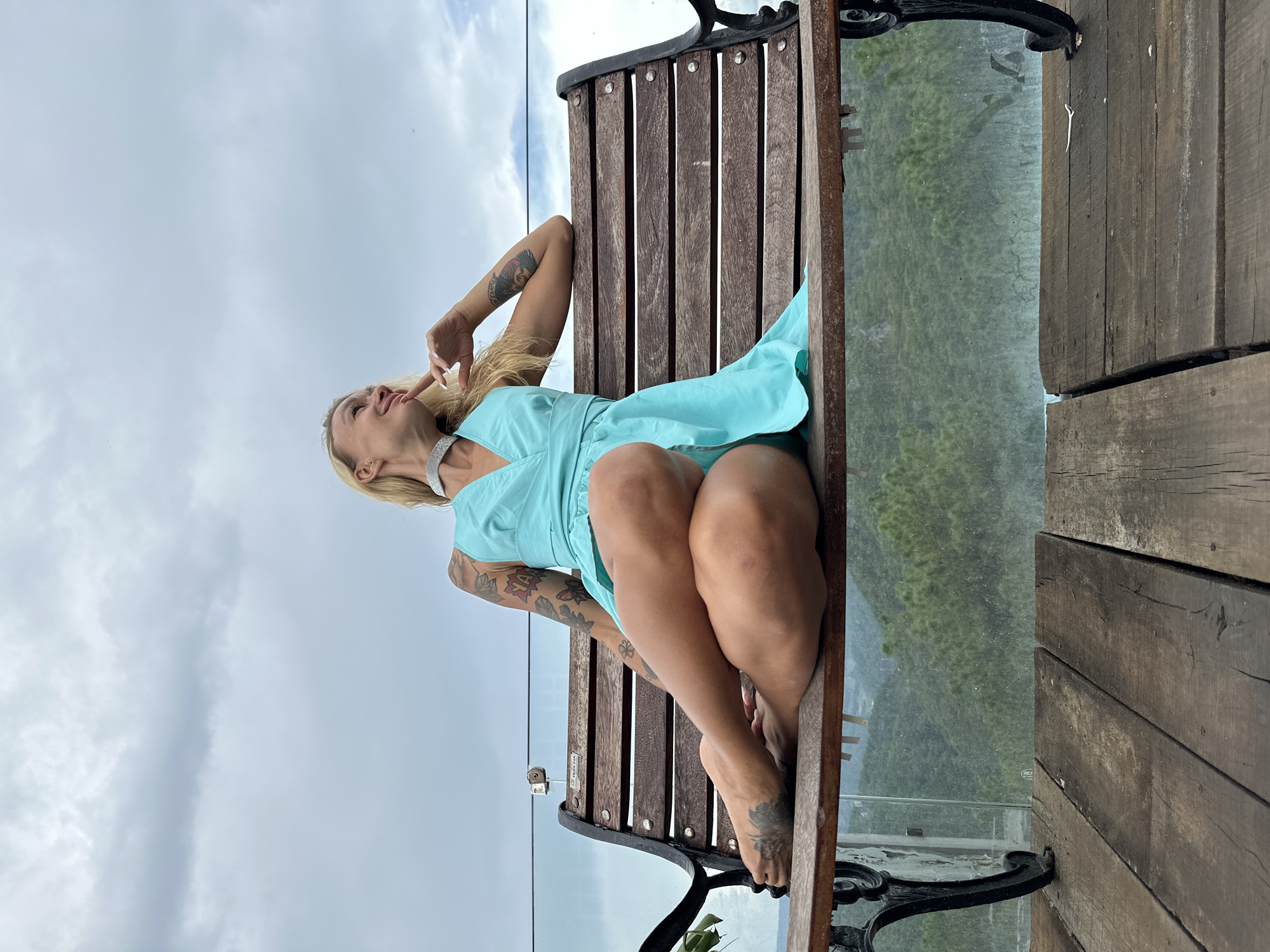 The image features a woman with blonde hair and tattoos sitting on a wooden bench outdoors. She is wearing a teal dress and a sparkly choker necklace. Her legs are crossed, and she is looking upwards with a slight smile. Behind her, there is a glass railing and a view of lush green hills under a cloudy sky. The bench is made of dark wood slats and has ornate black metal legs. The ground to the right of the bench is made of weathered wooden planks. The overall mood is relaxed and serene. The location appears to be a scenic viewpoint, possibly in Da Lat, Vietnam, given the lush mountainous terrain and cloudy weather typical of the region. There is no visible text in the image.