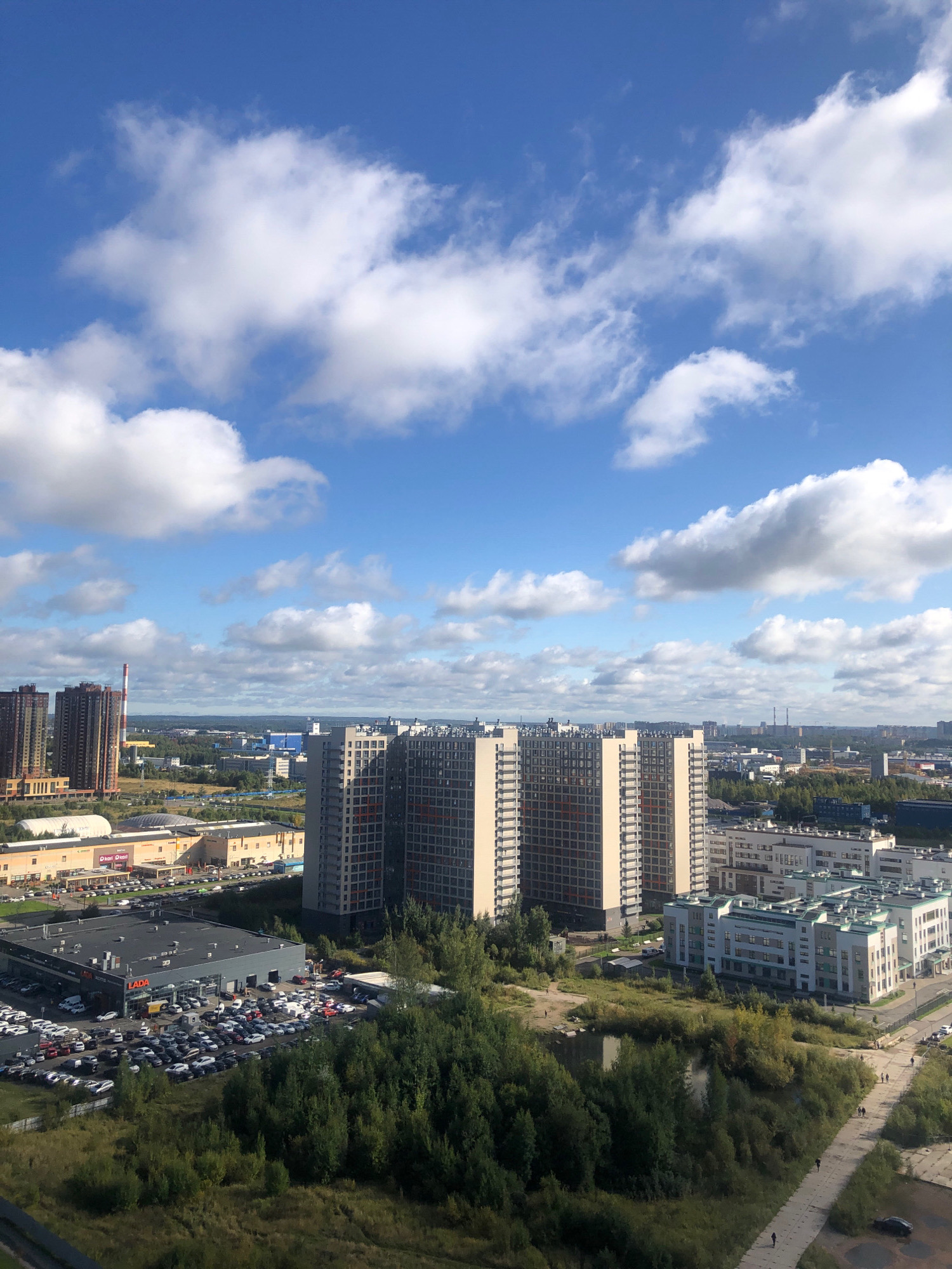 An aerial view of a city area in Novoe Devyatkino, Russia, is depicted on a clear day with blue skies and scattered white clouds. The scene features a mix of residential buildings, commercial structures, and green spaces.
Dominating the foreground and mid-ground are several large, multi-story residential buildings with visible windows and architectural details. To the left, a cluster of tall, dark-colored buildings stands out. In the center, a prominent, light-colored apartment complex with numerous windows is visible. Further to the right, a modern, white building with green accents appears to be a school or community center.
Below the residential complexes, commercial areas are evident. A large parking lot is filled with numerous cars, and a building with a sign that reads "LADA" suggests a car dealership. Another large building with a curved roof and what appears to be a shopping center with multiple entrances is situated in the mid-ground.
Green spaces are interspersed throughout the urban landscape. Dense trees form a significant part of the scene, particularly in the areas surrounding the buildings and along the edges of the developed areas. A small body of water, possibly a pond or lake, is visible amidst the trees in the lower right quadrant. A paved pathway runs alongside the trees, with several individuals walking on it.
In the background, more buildings and industrial structures can be seen, along with a tall smokestack with red and white stripes, a common feature in industrial zones. The overall impression is of a developing urban area with a blend of modern housing, commerce, and natural elements.