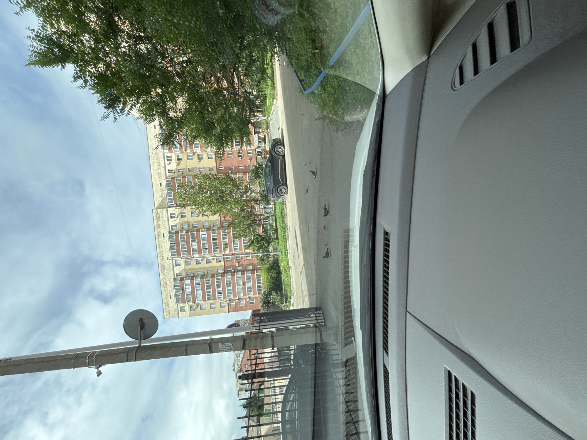 The image shows a view from inside a vehicle, looking out at an urban residential area. A tall, multi-story apartment building with a cream and red facade dominates the background. The building has many windows, and several air conditioning units are visible. In front of the building, there is a dark gray SUV parked on a paved area. Several pigeons are scattered on the ground in front of the car. A metal fence runs along the left side of the scene, and a concrete utility pole with a circular mirror attached stands near the fence. Lush green trees are present on the right side of the image. The sky is partially cloudy. This scene is consistent with a location in the city of Blagoveshchensk, Russia, a common urban environment with similar architecture and greenery.