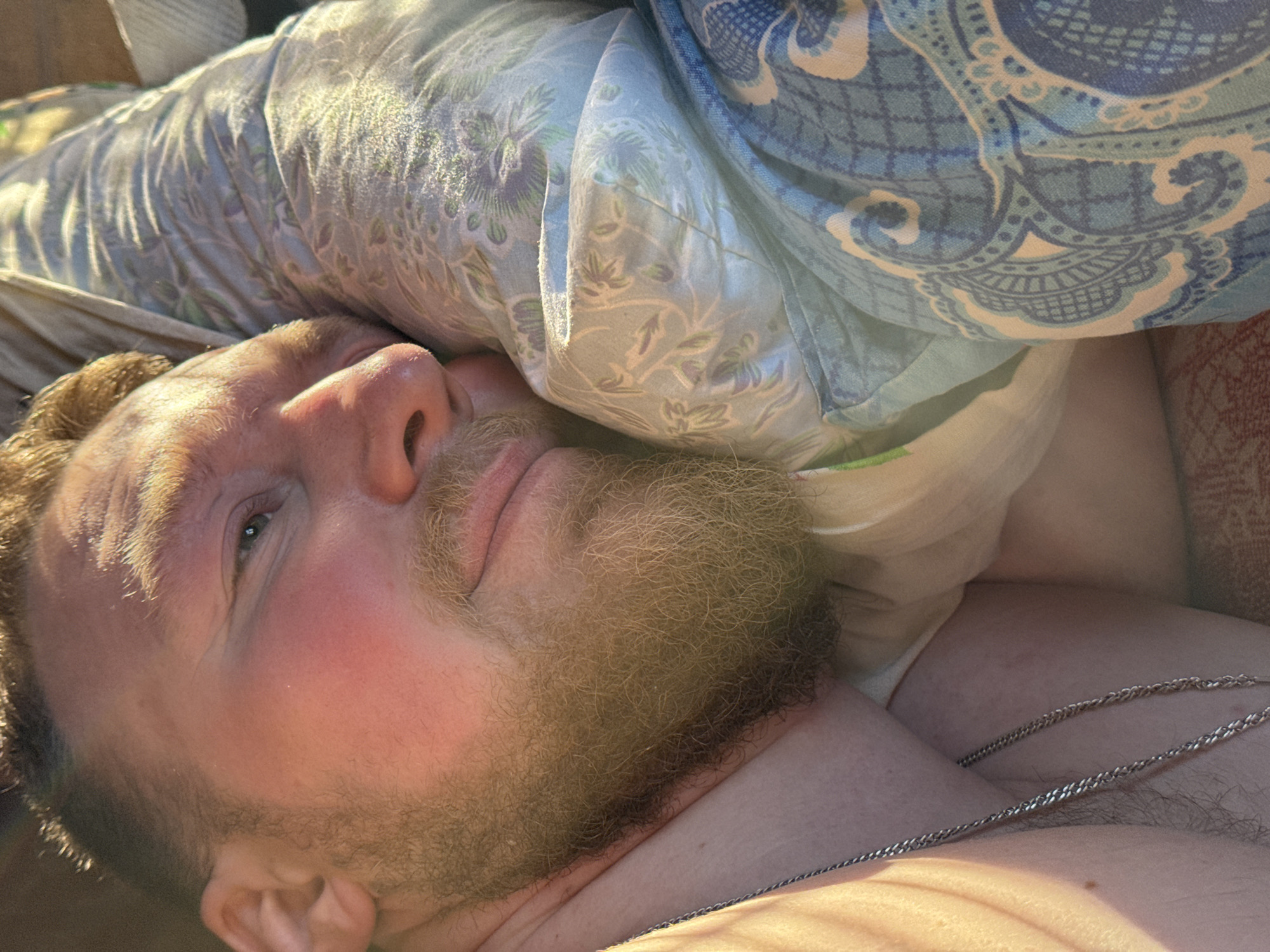 A Caucasian male is lying down, his head resting on patterned bedding. He has a reddish complexion, a full beard, and is wearing a silver chain around his neck. The background includes more patterned bedding. There is no indication of the location being Nizhnyaya Chermoda, Russia.
