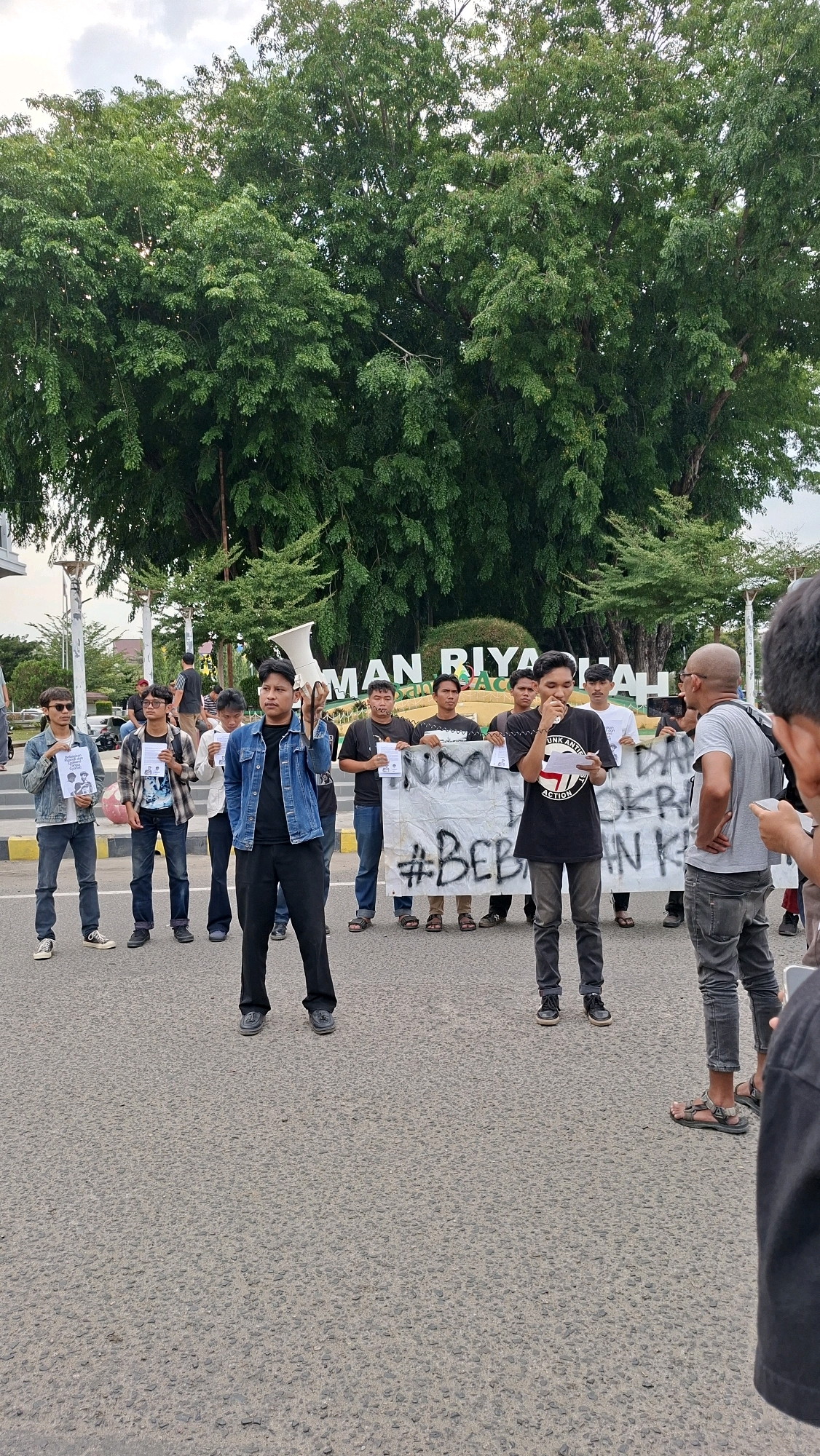 In Lhokseumawe, Indonesia, a group of young men are gathered outdoors, seemingly in a public space with a large, leafy tree and a sign visible in the background. The sign appears to read "UMAN RIYA". A banner with text in Indonesian and the hashtag "#BEBAS" is held by some of the individuals. One man in a denim jacket is holding a megaphone, and another man in a black t-shirt with a red logo is speaking into a smaller megaphone. Several individuals are holding white papers. The scene suggests a protest or demonstration. The ground is paved with asphalt, and the sky is overcast.