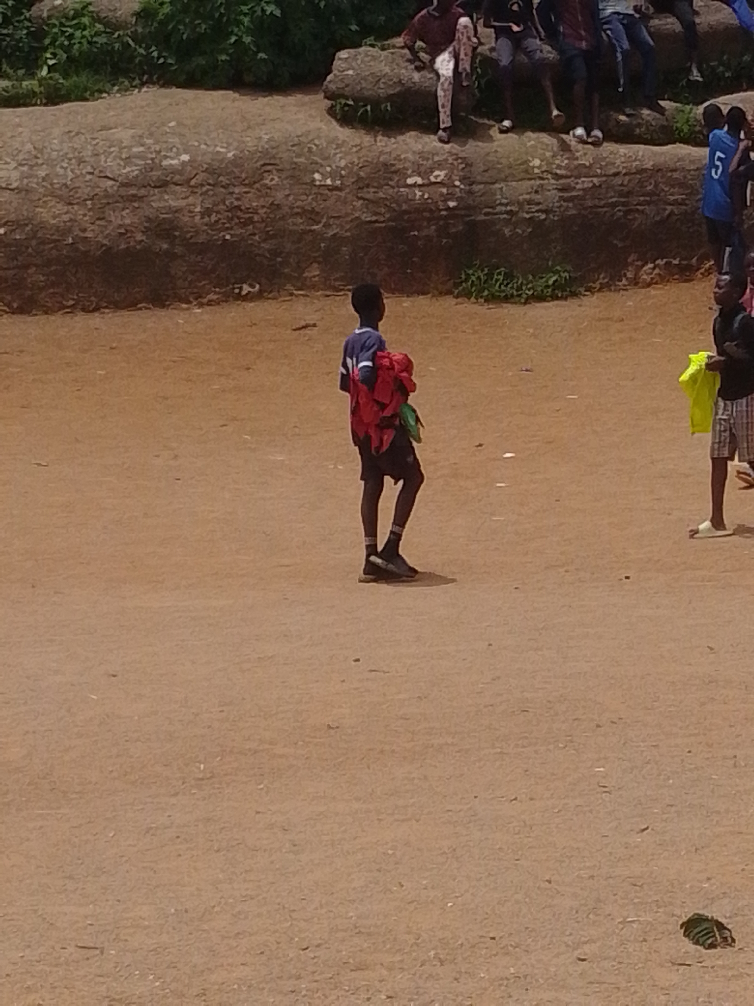 A lone boy stands in the middle of a dirt field, holding a bundle of red and green fabric. He is wearing a blue and white striped shirt and dark shorts. In the background, several other children and adults are seated on a large, rocky ledge. Some of the children are wearing sports jerseys. The environment appears to be an outdoor public space, possibly a park or a recreation area.