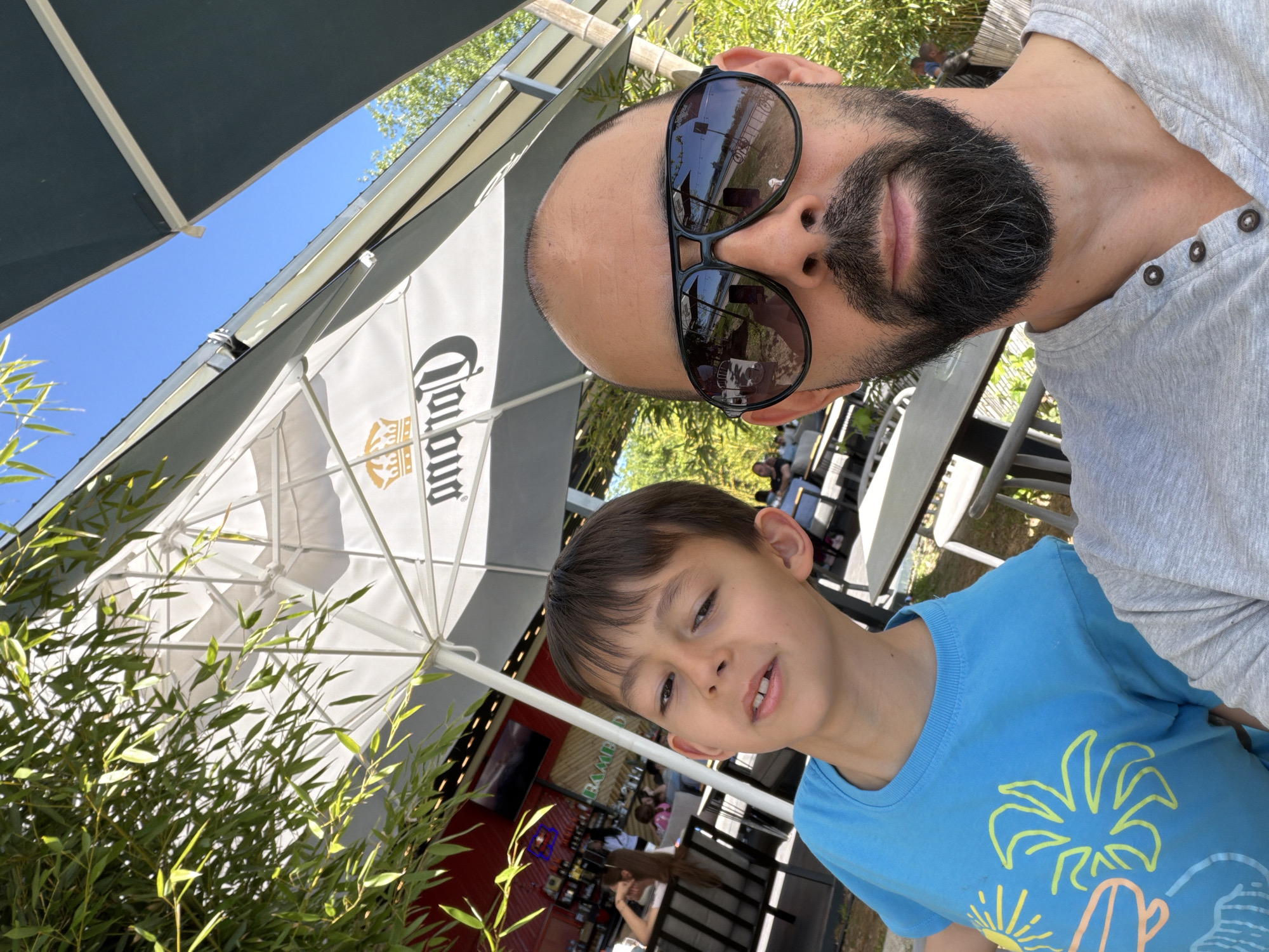 A selfie taken by a man with a beard and sunglasses features a young boy in a blue shirt with a palm tree design. They are outdoors, likely at a restaurant with a Corona-branded umbrella visible in the background. The image includes outdoor seating and some foliage. It is difficult to confirm this location is in Plovdiv, Bulgaria without additional evidence.