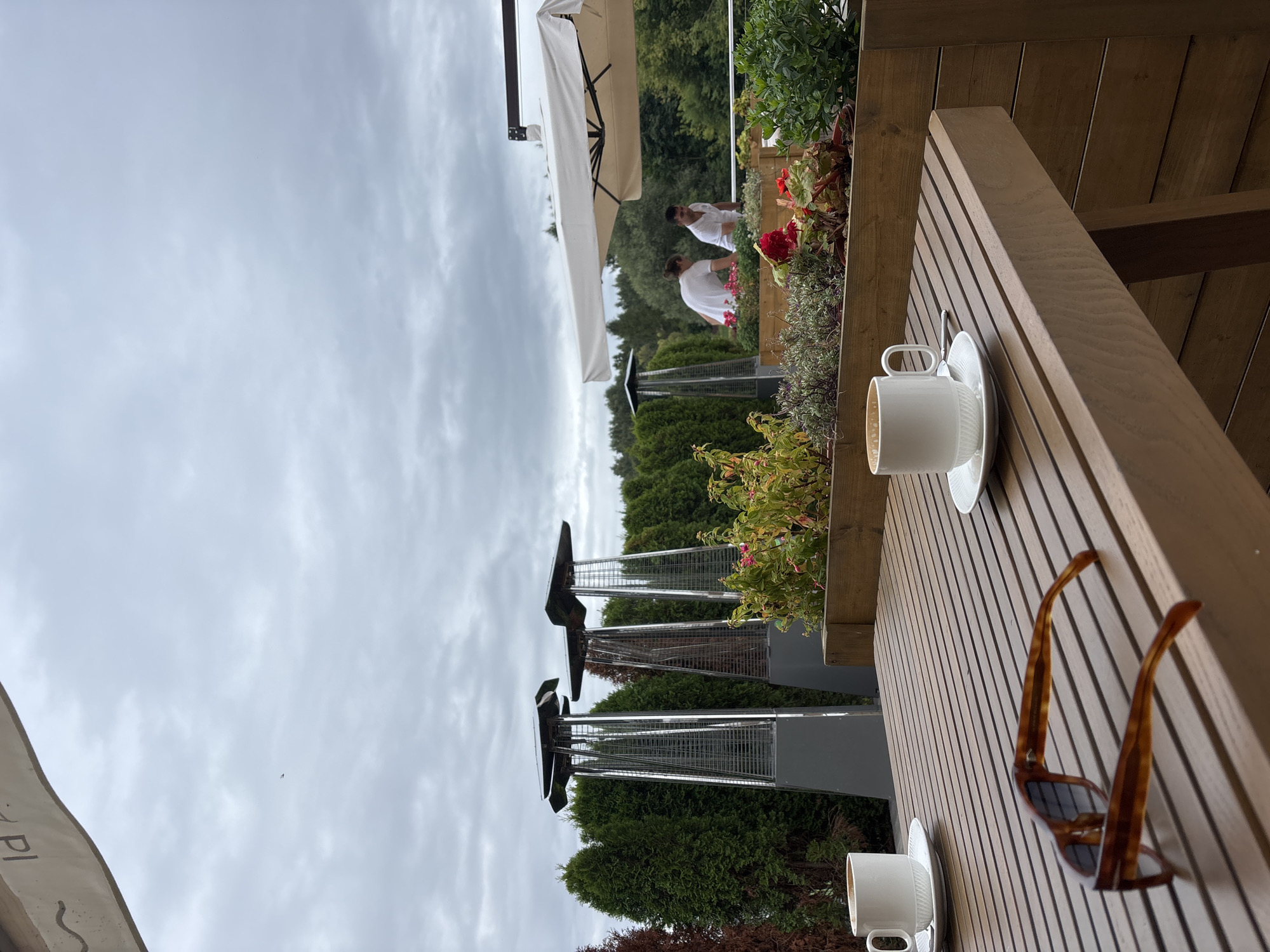 On an overcast day in Zvenigorod, Russia, a wooden table sits on an outdoor patio, set for two. Two white coffee cups and saucers are present. One cup is to the left, and another to the right. A pair of tortoiseshell sunglasses rests on the table in the foreground.
Behind the table, a planter box filled with green plants and red flowers runs along the edge of the patio. Two tall, silver patio heaters stand in the background, positioned between rows of tall green cypress trees. Further back, two individuals, dressed in white t-shirts, are tending to the plants in the planter box. They appear to be employees or staff members. Above them, a white umbrella provides shade. The scene suggests a restaurant or cafe with outdoor seating.