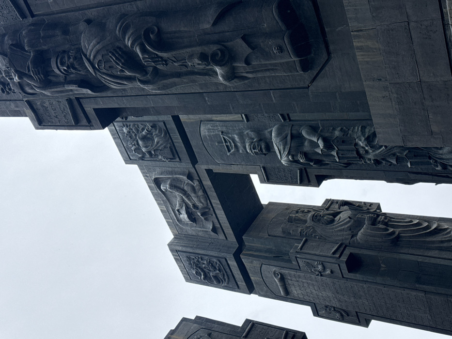 The image displays a large section of the Chronicle of Georgia monument, situated near the Tbilisi Sea in Tbilisi, Georgia. The structure comprises monumental dark grey stone pillars, some configured as arched gateways, extending upwards against an overcast white sky. The pillars are extensively covered with high-relief sculptures and larger-than-life full-figure statues. Depicted figures include bearded men in historical or religious attire, often holding objects such as scrolls, books, or miniature architectural models, including fortified structures and churches. Bas-relief panels illustrate narrative scenes, one clearly showing an individual kneeling and writing on a large sheet, while others depict groups of people and symbolic elements. Sections of Georgian script are inscribed on various surfaces of the monument. The perspective is upward-looking, emphasizing the colossal scale and intricate sculptural detail of the monument.