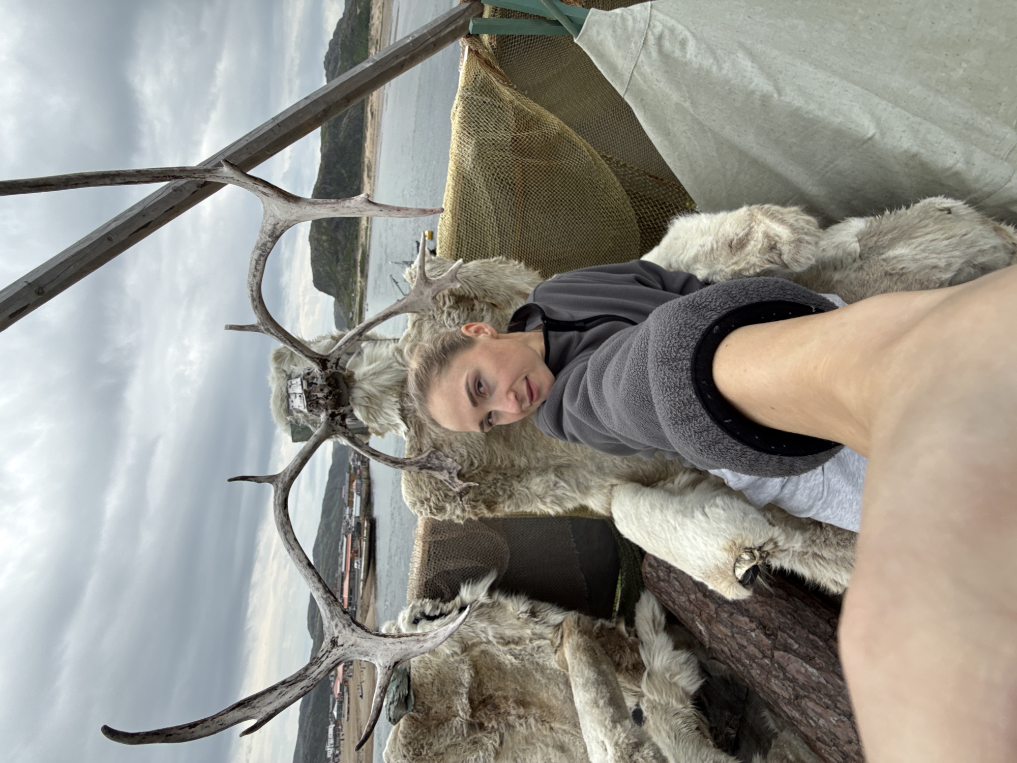 A woman is seated in a furry, rustic chair that appears to be made of reindeer hides and antlers. The chair is set up outdoors, with a body of water and a coastal town visible in the background. The town, Teriberka, Russia, features a cluster of buildings near the shore, with hills rising behind it. The sky is overcast with thick clouds. The woman is taking a selfie, with her arm and hand extended into the foreground. She is wearing a grey hooded jacket and grey sweatpants. The chair itself is elaborate, with large reindeer antlers forming a backdrop behind her head. Some wooden beams and netting are also part of the structure she is sitting in.