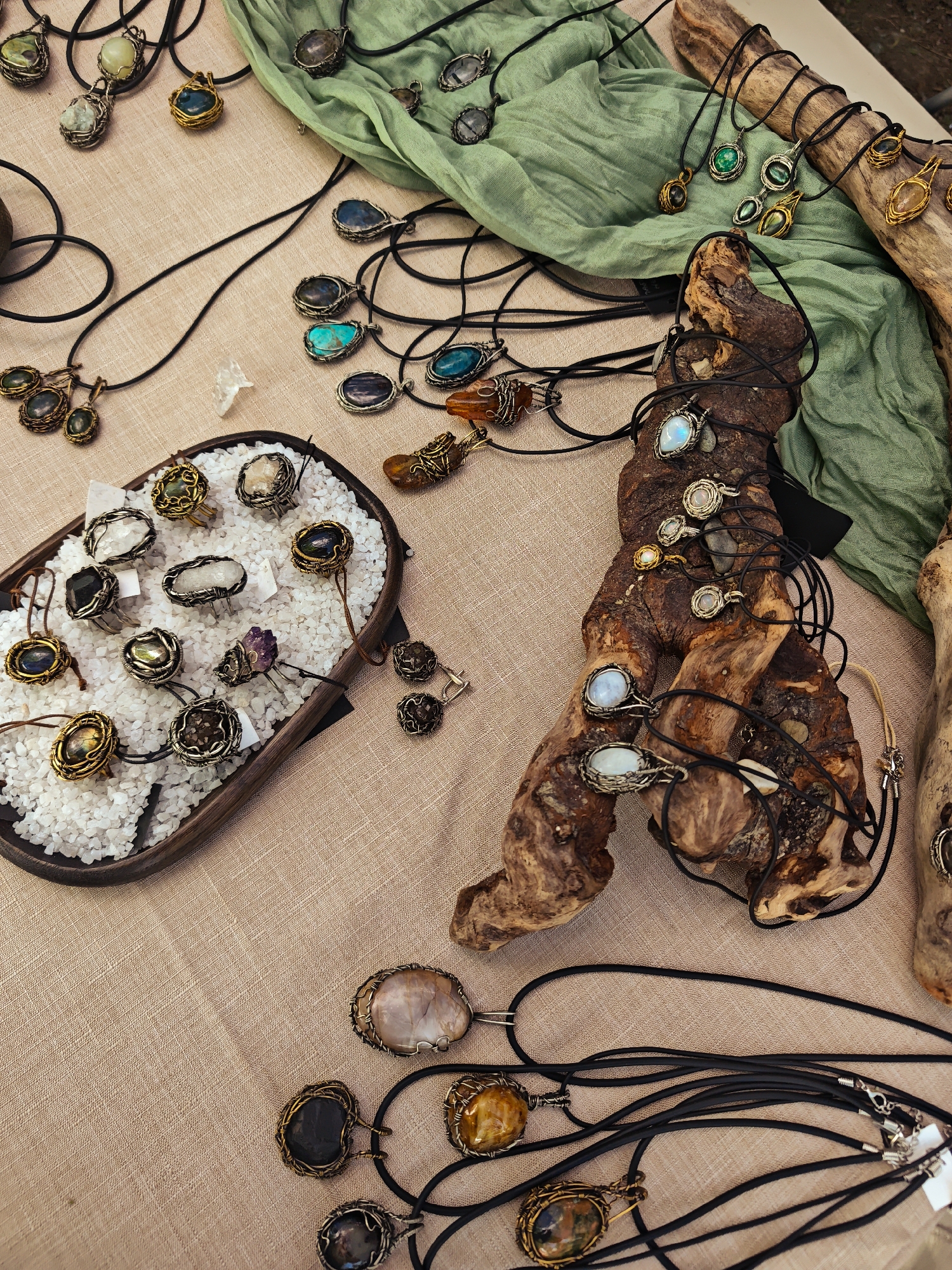 A top-down, medium shot captures an outdoor market stall with an assortment of handmade jewelry displayed on a textured, tan-colored fabric. The primary focus is on various necklaces and rings, predominantly featuring natural stones and crystals encased in wire-wrapping.
In the center-left, a dark wooden tray is filled with small white pebbles, serving as a base for several rings. These rings showcase stones like amethyst, clear quartz, and various opaque gems. To the right of the tray, a gnarled, light brown piece of driftwood serves as a rustic display for multiple necklaces. These necklaces feature pendants with moonstone, opal, and other iridescent stones. Further to the right, a green, crinkled fabric is draped, with more necklaces adorned with blue, green, and amber-colored stones. A few additional necklaces with dark stones and amber pendants are scattered on the tan fabric in the foreground. The overall aesthetic suggests a bohemian or naturalistic style of craftsmanship. There are no people visible in the image, and the setting appears to be a market or fair, though no specific identifying landmarks for Kimry, Russia, are present in the visual information.