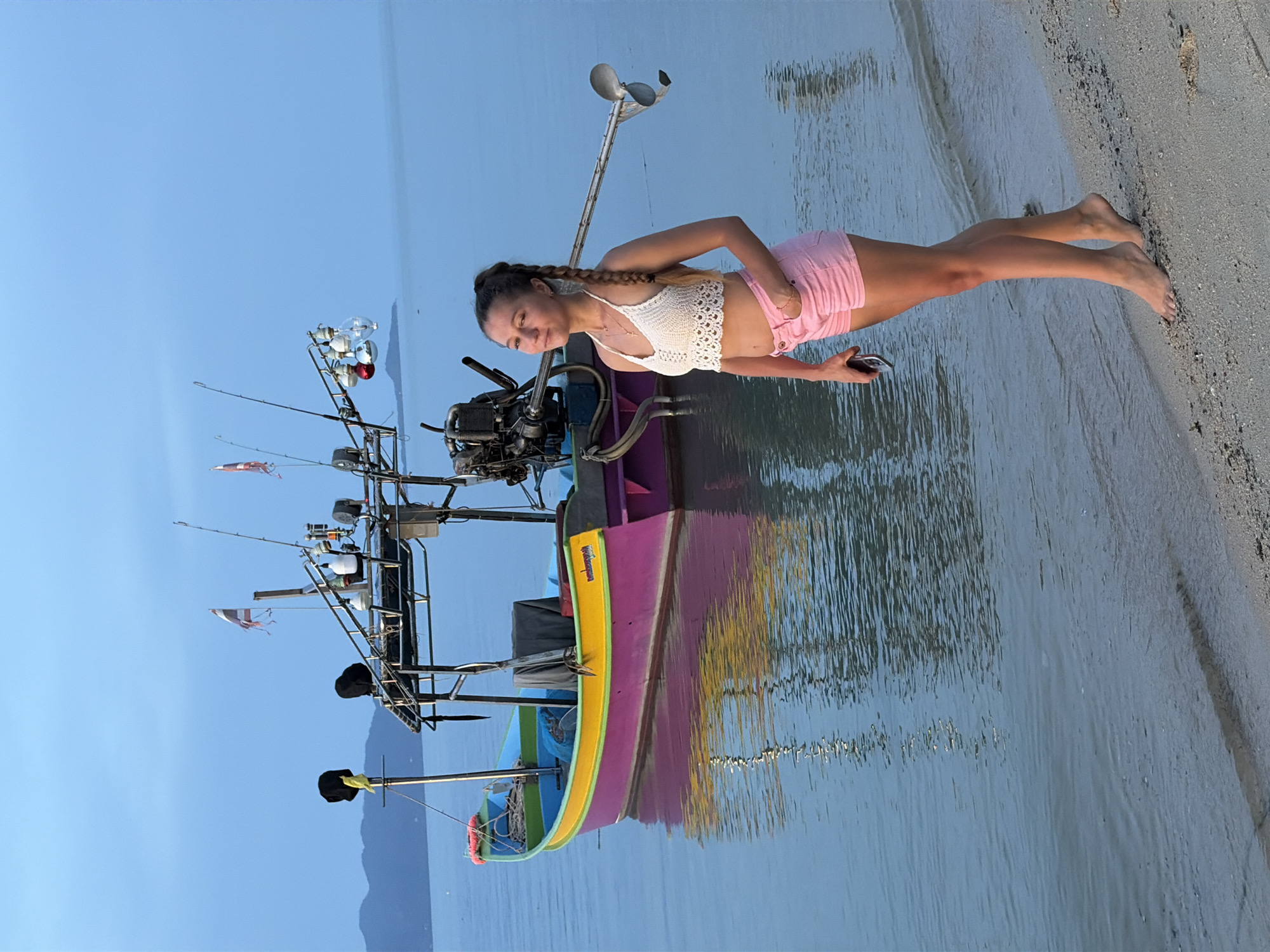 This media file depicts a young woman with braided hair standing on the shore of a body of water, with a colorful fishing boat partially submerged nearby. She is wearing a white crochet top and pink shorts. Her right arm is extended, and she appears to be holding a mobile phone. The boat is a prominent object in the scene, featuring a multi-colored hull in shades of purple, yellow, and blue, with a motor at the back and fishing equipment such as rods and tackle visible.
The scene is set on a sandy beach with calm, reflective water. The time of day appears to be either early morning or late afternoon, judging by the soft, diffused light and the calm water, suggesting a tranquil atmosphere. The sky is a clear, pale blue. The location is identified as Baan Thong Po, Thailand, which suggests a tropical or subtropical setting.
There is some visible text on the side of the boat, though it is not entirely legible due to the image's orientation and resolution. The woman's posture and the relaxed setting imply a moment of leisure or preparation, perhaps for fishing or simply enjoying the scenery. The overall mood is peaceful and serene.