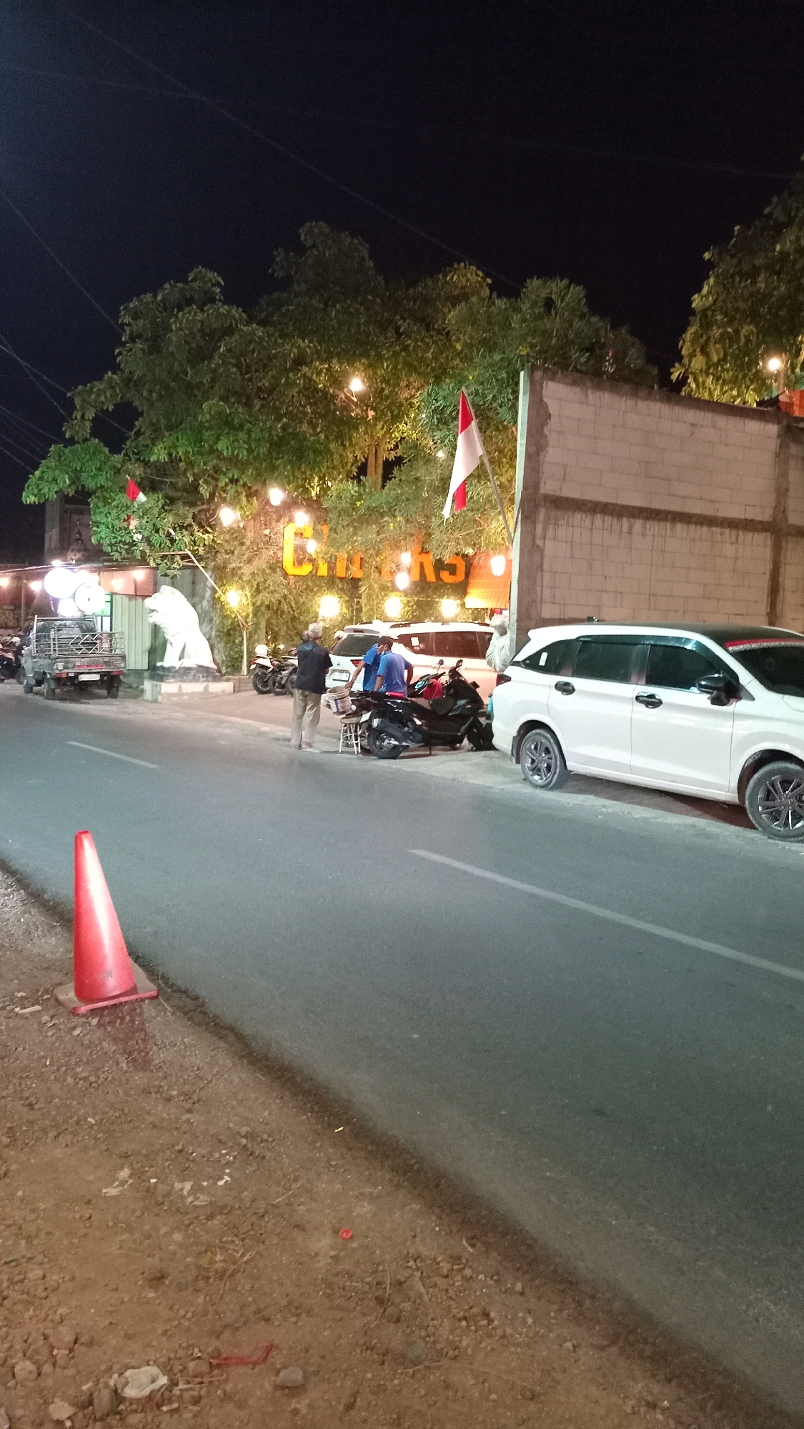 An exterior, nighttime, medium shot shows a street scene in Suci, Indonesia. On the left, a red traffic cone is positioned on the gravel shoulder. The street itself is paved and appears to be a two-lane road. In the background, a building with a brick facade is visible. Trees with fairy lights strung through them line the front of the building, creating a festive atmosphere. A large white sculpture of a lion stands in front of the building, near a small pickup truck. Several motorcycles and two white vans are parked in front of the building. Two men are standing near the motorcycles, and one appears to be interacting with a third man who is sitting. A white SUV is parked on the right side of the street, facing away from the camera. The overall impression is that of a lively street-side establishment.