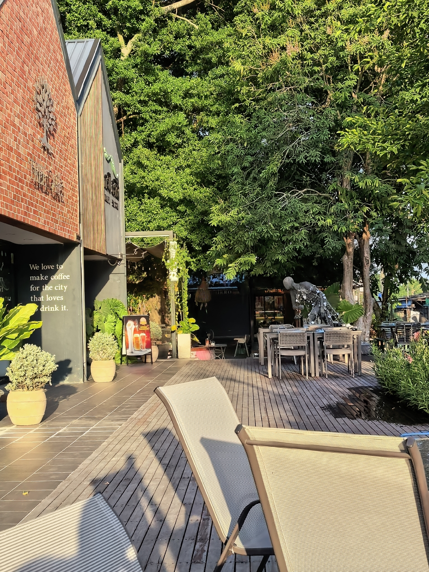 This image depicts the outdoor seating area of a cafe, possibly "The Coffee Shop" as suggested by the signage, located in the city of Ko Samui District, Thailand. The cafe features a modern architectural design with a prominent brick facade and a sloped roof. In the foreground, two lounge chairs with striped cushions are positioned on a wooden deck. The deck extends towards a shaded area where several tables and chairs are set up. A striking metal sculpture of a person stooping over is placed on one of the tables. Lush green trees and foliage surround the seating area, creating a serene and natural ambiance. In the background, a glimpse of other establishments and tropical vegetation is visible, indicating a vibrant and lively environment. The scene is bathed in warm, natural light, suggesting it was taken during the late afternoon or early morning.