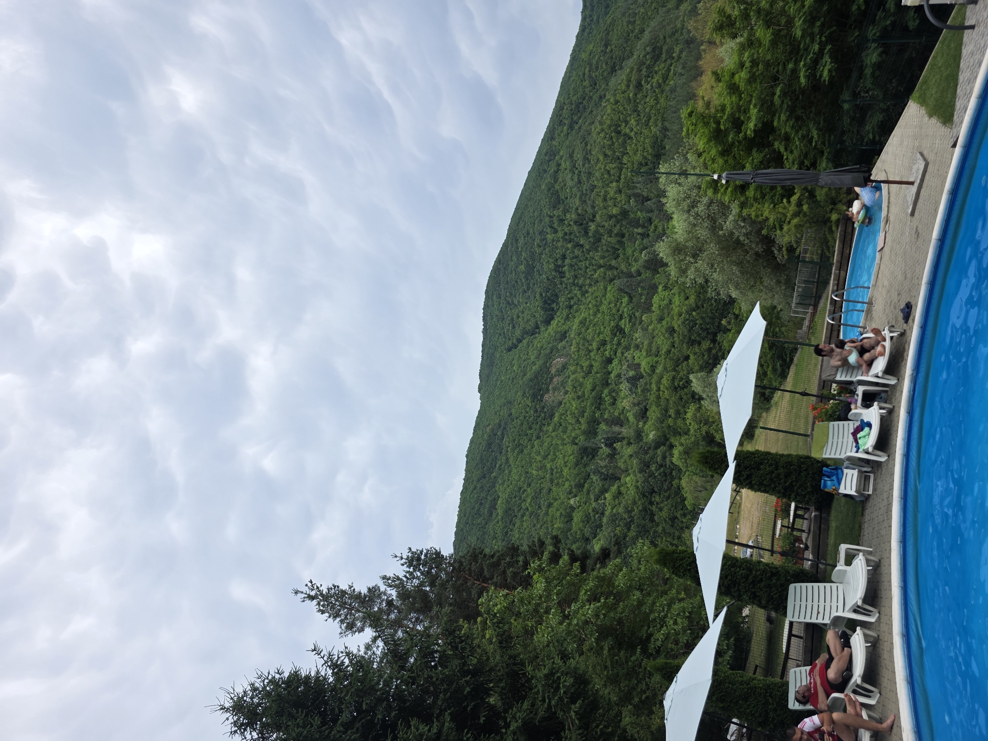 The image depicts an outdoor scene at a resort or private residence in Lukovo, Bulgaria, featuring two swimming pools. In the foreground, a bright blue swimming pool is partially visible, with its edge and a section of its water surface taking up the bottom left quadrant of the frame. Adjacent to this pool, a paved area is lined with several white lounge chairs, some occupied by people. Three white umbrellas are set up over some of these chairs.
To the right, a second, smaller swimming pool with blue water is situated on a slightly elevated, paved area. A few people are in or near this pool; one man is sitting in a lounge chair holding a child, while another person is in the water with an inflatable. A dark umbrella is closed and stands on a pole near this pool.
Behind the lounge area, lush green trees and cypress-like shrubs are visible. Beyond the immediate vicinity of the pools, a steeply sloping, densely forested mountainside dominates the background, with a cloudy, grey sky overhead. The overall impression is one of a recreational area set against a natural, verdant landscape.