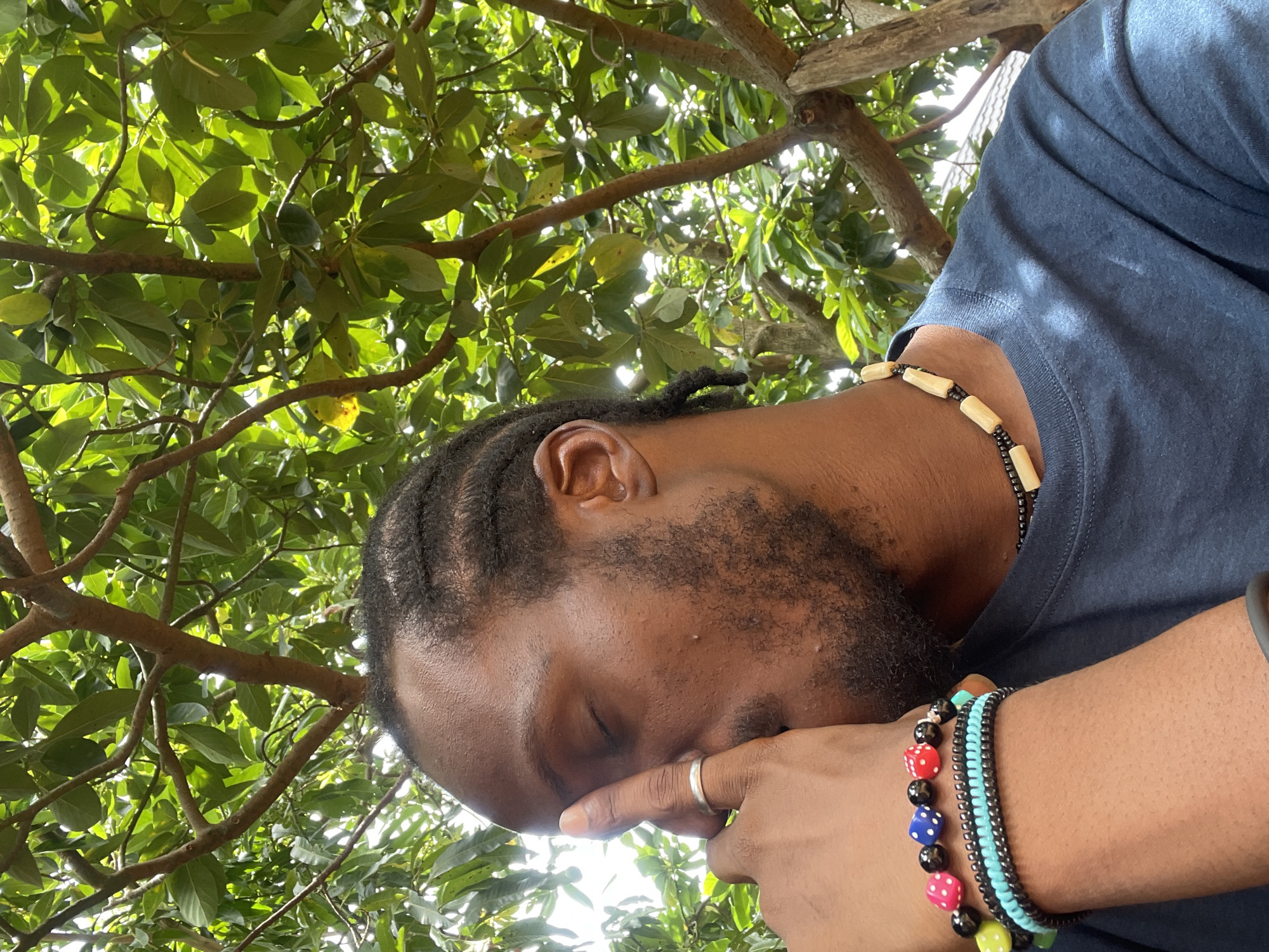 A medium shot taken from a low angle depicts a Black male with dreadlocks and a beard. He is wearing a blue shirt and several bracelets, including one with colorful dice beads. He has a ring on his finger and a beaded necklace around his neck. He appears to be holding his face, with his eyes closed. The background consists of a tree with green leaves and branches. The location is Jos, Nigeria, based on the presence of tree species common in the area and the ethnicity of the person pictured.