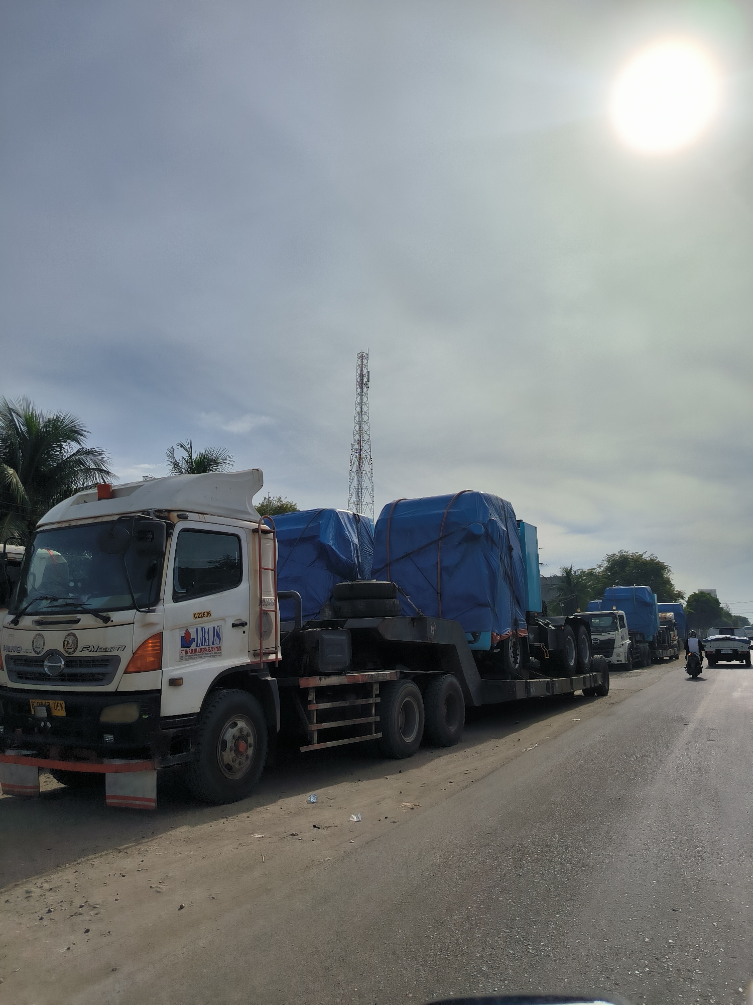 This image shows a procession of trucks carrying large, blue-tarped loads on a paved road. The trucks are lined up in a convoy, suggesting a transport operation. The lead truck, a white Hino, is clearly visible in the foreground, with its company logo and vehicle identification number visible on the side. The sky is bright and overcast with a sun partially visible, indicating daylight. There are palm trees and a tall transmission tower in the background, suggesting a semi-tropical or coastal environment. A person on a motorcycle is visible in the distance, traveling in the same direction as the trucks. The road appears to be a two-lane highway with a dirt shoulder. The scene is likely located in or near the city of Tingkeummanyang, Indonesia, given the context provided.