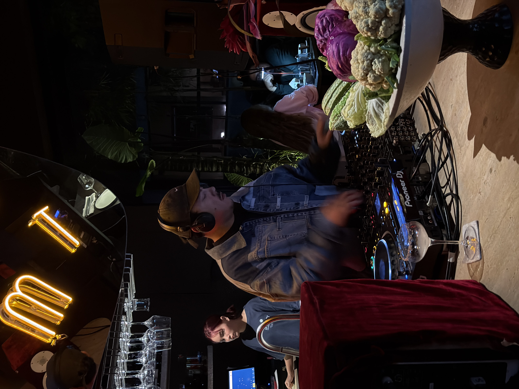 The image captures an indoor event, likely a party or gathering, set in Mengwi, Indonesia. The scene is dimly lit, suggesting it's evening or night. In the foreground, a person wearing headphones and a denim jacket is seen operating a DJ console, indicated by the presence of a Pioneer DJ mixer and turntables. Their arm is blurred, suggesting movement, and they appear focused on the task. Behind the DJ, a large decorative arrangement of vegetables, including cauliflower and red cabbage, is prominently displayed.
Further back, other individuals are visible. One person is partially obscured, and another, with red hair, is seated at a table with a computer monitor. The overall atmosphere seems lively and engaging. Yellow neon signs spelling out "T" and "m" are visible on a reflective surface, adding a modern, urban touch to the setting. The presence of wine glasses on a rack also suggests a celebratory occasion. The interior appears to be a contemporary space with a mix of modern and artistic elements, possibly a restaurant, bar, or event venue.
