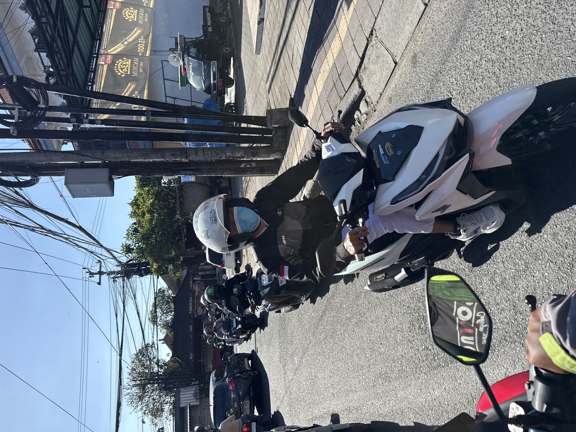 In Denpasar, Indonesia, a line of motorcyclists navigates a sunlit street on a clear day. The foreground features a person on a white scooter, wearing a white helmet, light blue mask, and a dark jacket with white accents. They are positioned behind a motorcycle with the license plate "DK 1250 WR". In the middle ground, other motorcyclists are visible, including one in a green helmet and another on a dark scooter. Further back, a black pickup truck with "PATROLI" and "KEL PEDUNGAN" written on it is parked on the right side of the road. The street is paved and lined with buildings and utility poles with numerous cables. Lush green trees are interspersed along the sides of the road. The overall scene depicts a typical urban traffic environment in Denpasar.