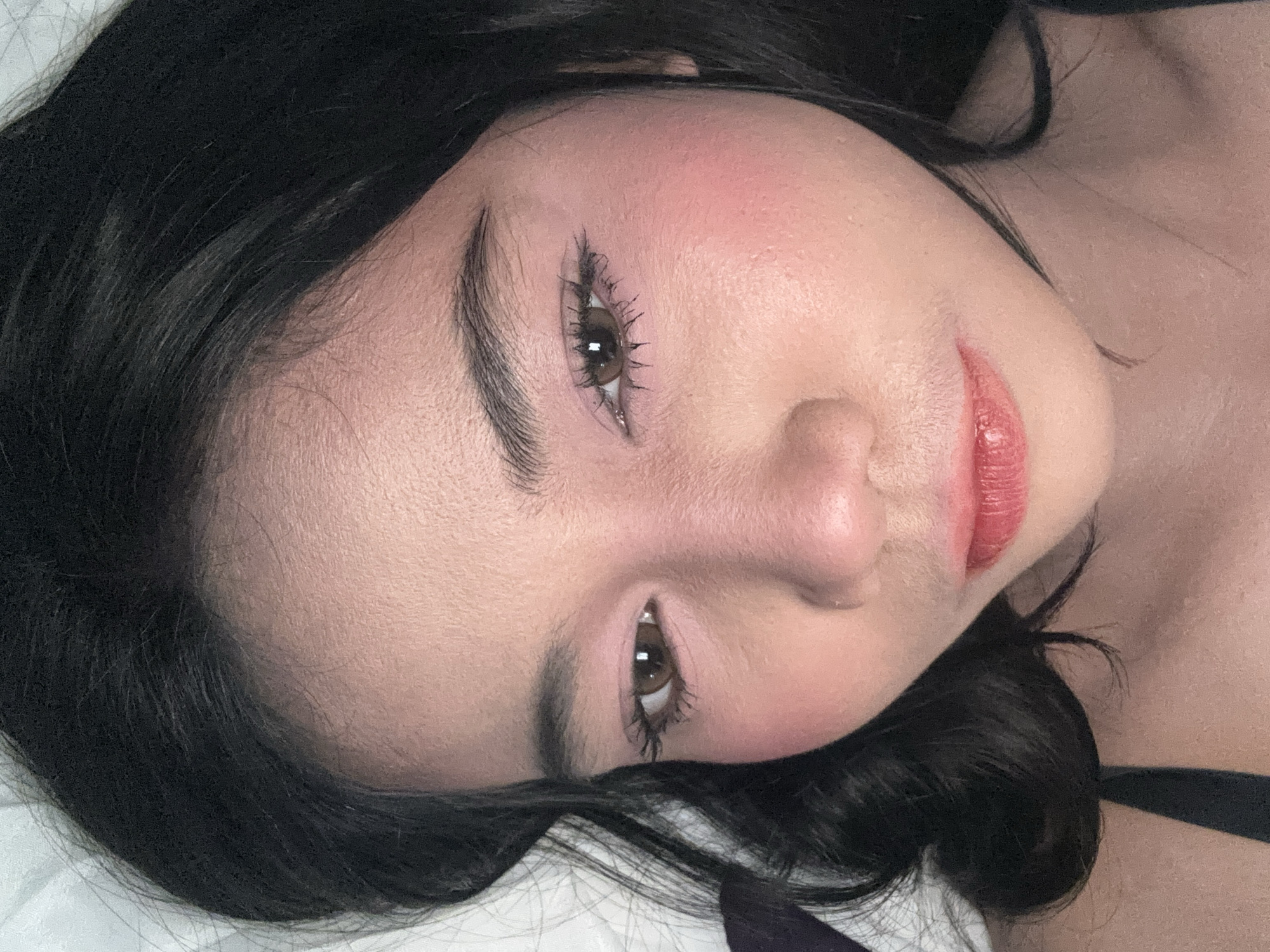 A close-up, high-angle portrait features an adult female situated in North Jakarta, Indonesia. The subject has dark, wavy hair that frames her face, and a light-to-medium skin tone exhibiting visible texture and fine facial hair. Her dark brown eyes are slightly downcast, revealing pink eyeshadow on the eyelids and dark mascara, with notable clumping on the lower lashes. A prominent application of reddish-pink blush covers her cheekbones and extends towards the bridge and tip of her nose. Her full lips are coated in a glossy red or deep pink lipstick. A dark, thin strap, consistent with an undergarment or top, is visible over her left shoulder (viewer's left). The background consists of soft, light-colored fabric, out of focus, likely depicting bedding or a pillow.
