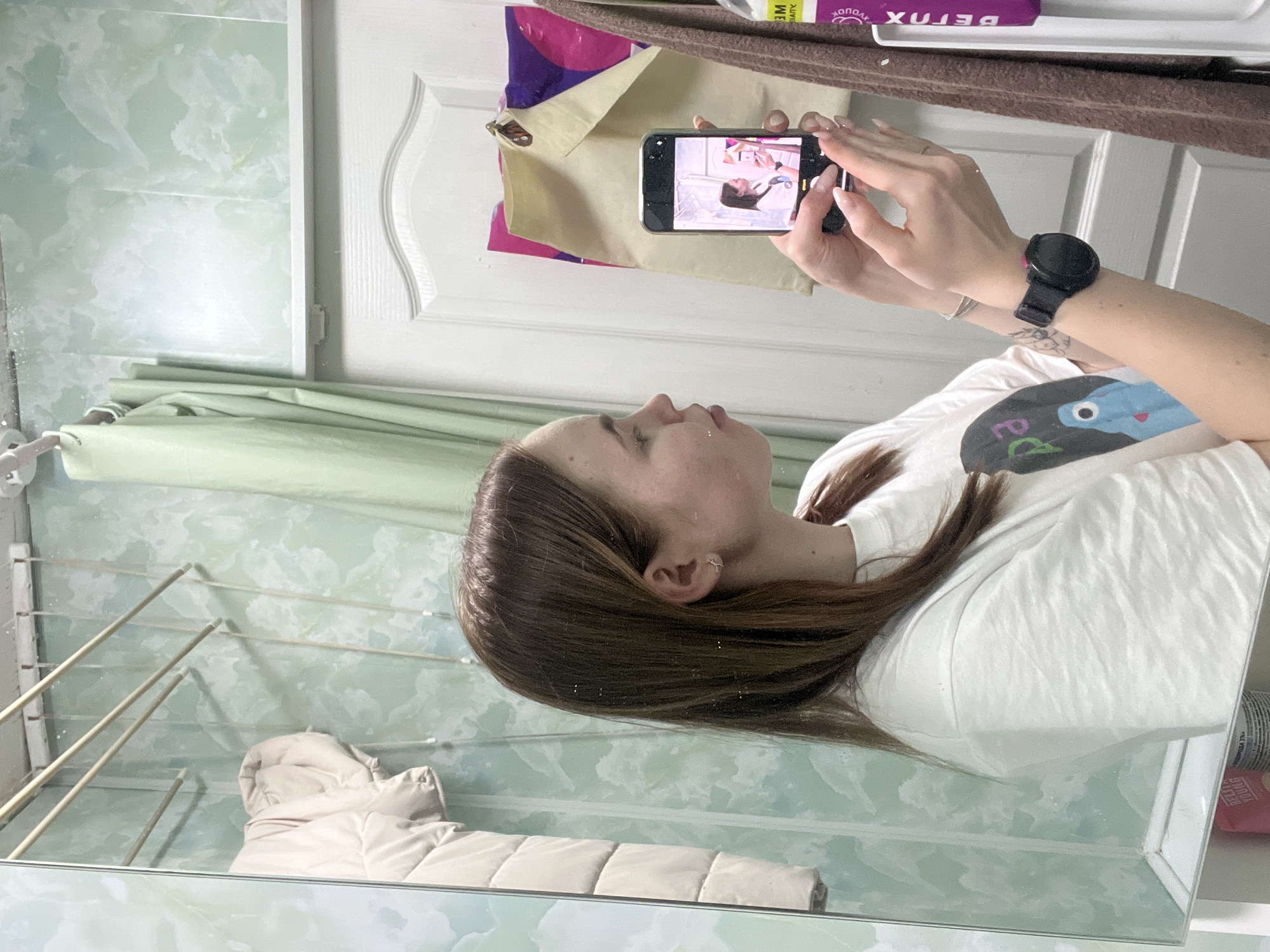 A woman is taking a photograph of her reflection in a mirror. The location appears to be a bathroom, identifiable by the tiled walls and shower curtain. A white coat is visible on a clothes rack near the mirror. The woman is wearing a white t-shirt and a black smartwatch on her left wrist. She is using a smartphone to capture the image. The mirror reflects a view of the room, including part of a white door with decorative panels. There are various items visible on a shelf behind the woman, including a purple tube with Russian text. The photo does not confirm that this is Chaikovsky, Russia.