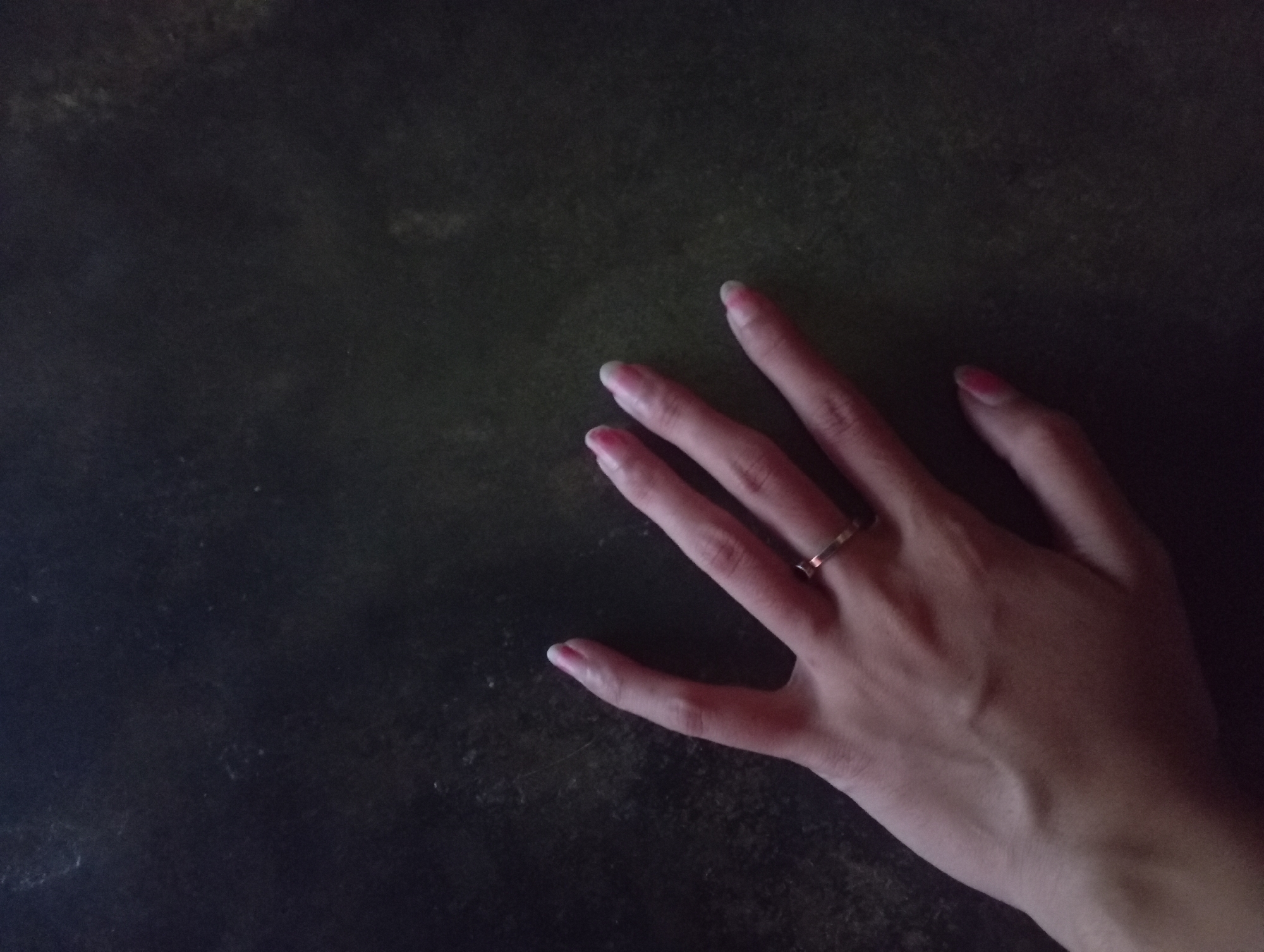 A left hand, with a thin gold ring on the ring finger and painted fingernails, is positioned against a dark, textured surface. The hand is angled with the palm facing downwards and slightly towards the viewer. The fingertips are fanned out, with the index and middle fingers extended. The background is a dark, possibly wooden or stone surface, with subtle variations in tone and texture. There are no other people or objects visible in the frame. The scene is illuminated from an unseen source, creating shadows and highlighting the contours of the hand. The location in Rejosari, Indonesia, is not discernible from the visual elements of the image.