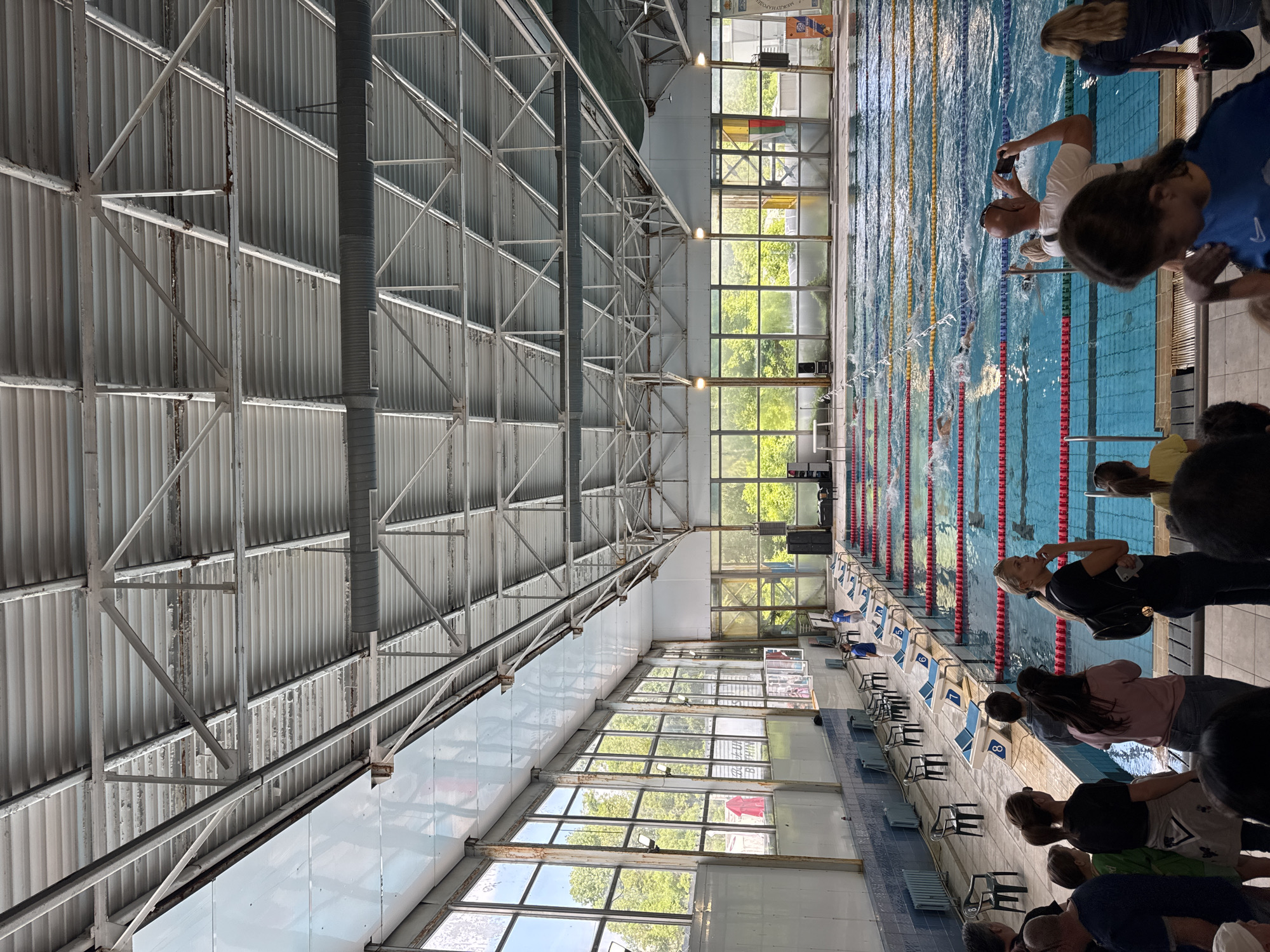 The image shows an indoor swimming pool during a swimming competition. Swimmers are in several lanes, actively swimming. Spectators, including a man taking pictures with his phone, are watching from the side. The pool has lane markers and starting blocks numbered 1 to 9. The pool area is surrounded by large windows. The structure of the building includes a complex metal framework for the roof. Based on the building’s design, this location is likely the swimming pool complex "Spartak" in Sofia, Bulgaria.