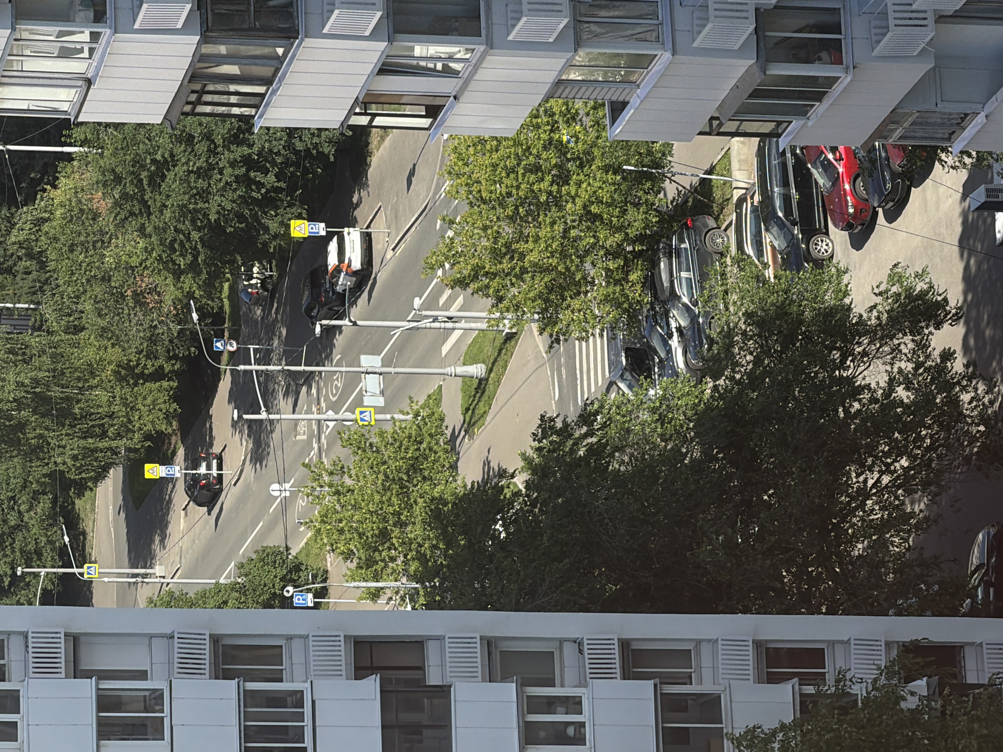 The image shows a view from a multi-story apartment building looking down at a street and parking area in the city of Khimki, Russia. The apartment building, a prominent white structure with balconies, occupies the left side of the frame. The street below is lined with trees and features several parked cars. Visible vehicles include a dark sedan, a white and orange car, and several other cars parked in a lot including a red SUV. There are pedestrian crossings marked on the road, and several traffic signs are visible, including parking signs and a speed limit sign. The environment is sunny with shadows cast by the trees and buildings. No people are clearly visible in the image.