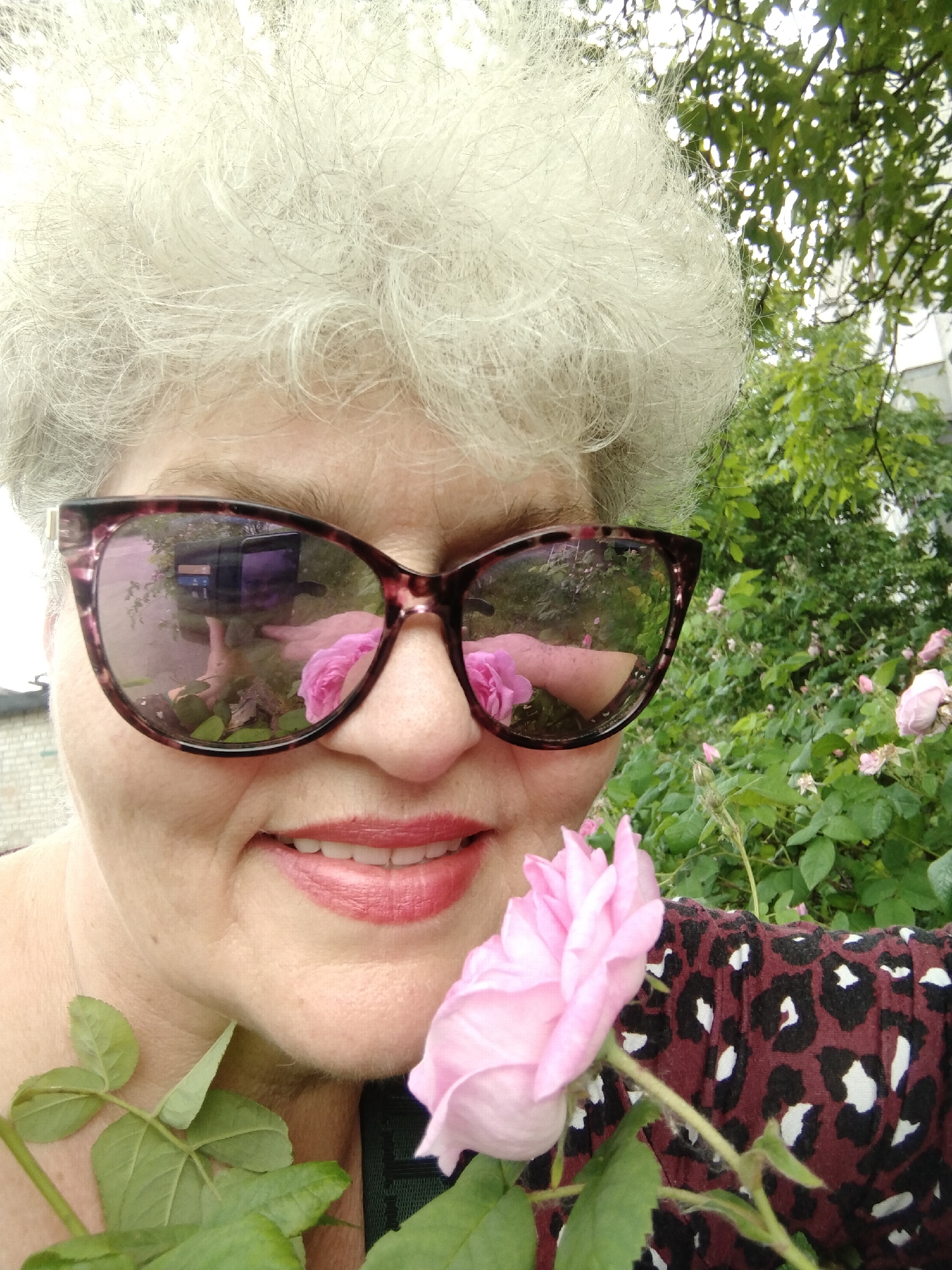A close-up, outdoor portrait features an older woman with short, curly, light grey hair, smiling gently at the viewer. She wears large, cat-eye style sunglasses with a dark tortoiseshell frame. The reflective lenses of her sunglasses prominently display a distorted reflection of her arm and hand holding a dark-colored smartphone or camera, suggesting she is taking a selfie. Also visible in the reflection are several pink roses and green foliage.
A vibrant light pink rose with green leaves is positioned in the foreground near her right shoulder, almost touching her chin. Her lips are painted with a rosy-pink lipstick, complementing the color of the rose. On her right side, a portion of her clothing is visible, featuring a maroon or burgundy background with a black and white leopard print pattern.
The background consists of lush green foliage, suggesting a garden or park setting, likely filled with rose bushes. Above and to the right, a bright sky is visible through the tree leaves. To the left, a light-colored building, possibly brick, can be faintly seen. The scene is brightly lit, indicating daytime. The woman's smile conveys a sense of happiness and enjoyment. The image captures a serene moment with nature in Horishni Plavni, Ukraine. There is no visible text in the image.