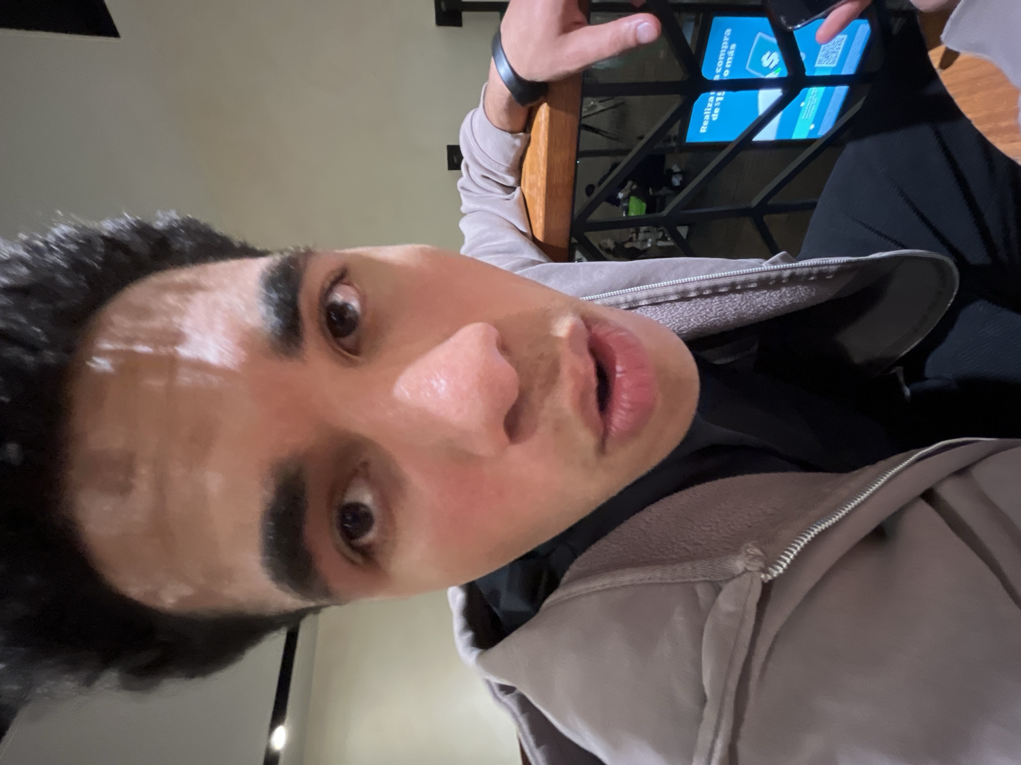 A low-angle selfie of a person with dark hair, dark eyebrows, and light skin, likely a young man, wearing a gray zippered jacket over a dark shirt. The person's face fills much of the frame, with the nose being prominent. The background is slightly blurred but shows a structure made of black metal bars, with a sign reading "Realiza tu compra o más", indicating a place where purchases can be made. The sign also contains a QR code. A hand is holding a smartphone over the black metal bars, suggesting the person might be scanning the code or using the phone for payment. Given the sign's wording in Spanish, it is likely taken in a Spanish-speaking location. The style of the metalwork in the background is often used in the city of Puebla, Mexico.