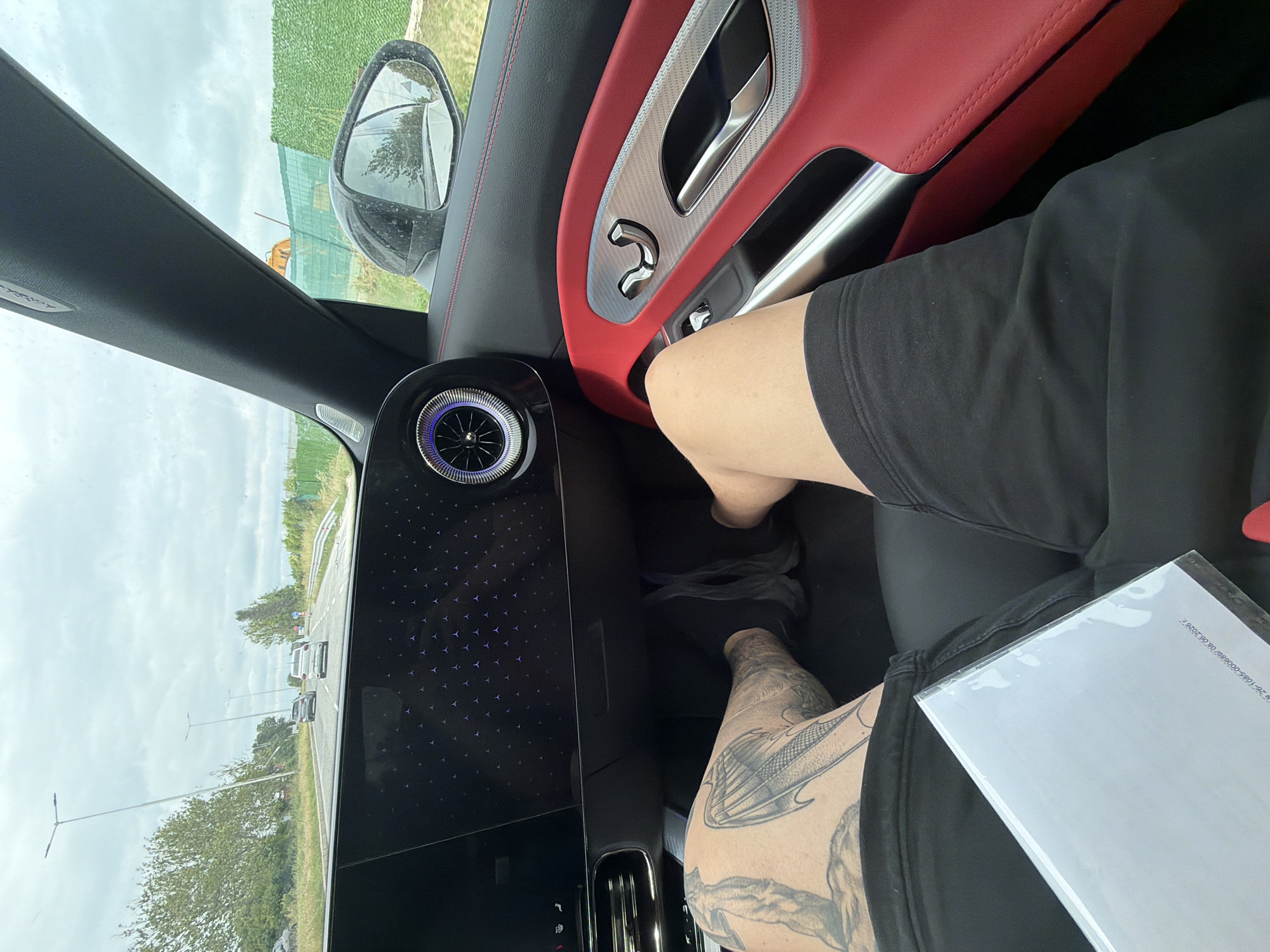 The image shows the interior of a car with the driver's perspective. The dashboard features a large, dark screen displaying a pattern of small, illuminated stars, and a circular air vent with blue illumination. The interior of the car has red leather accents on the door panel and seats. The driver's legs are visible in the foreground; the left leg has a prominent tattoo. The driver is wearing black shorts. A white paper object, possibly a document or a tablet, is held in the driver's lap. Outside the car, visible through the windshield, is a highway with cars driving, streetlights, trees, and a cloudy sky. To the right, a side mirror reflects greenery and some construction fencing. The location is identified as Dolno Ablanovo, Bulgaria.