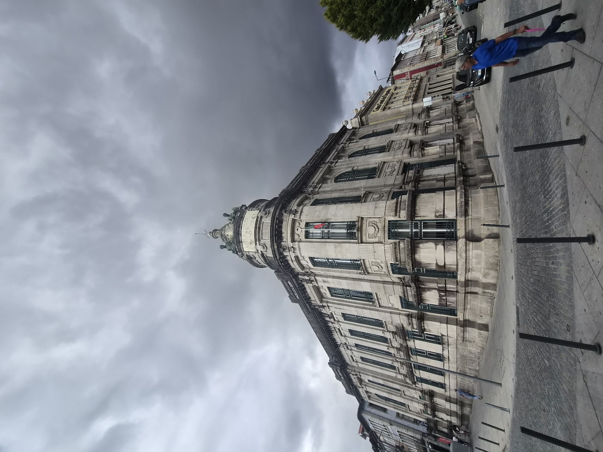 The image captures a street view in Braga, Portugal, dominated by a large, ornate stone building with a domed cupola. The architecture suggests a historical or civic building, possibly a bank or government office, with intricate stonework, tall windows, and decorative elements. A Portuguese flag is visible on one of the balconies. The sky is heavily overcast with dark, dramatic clouds, indicating impending rain or a stormy day.
In the foreground, a man wearing a blue shirt and jeans walks across the cobblestone sidewalk, carrying a pink object. His body is angled away from the camera, and he appears to be walking from right to left. The street is lined with bollards, and other buildings with visible signage are present in the background, suggesting a bustling urban environment. A few other pedestrians are visible further down the street, and vehicles are parked or moving on the road. The overall atmosphere is somber due to the weather, contrasting with the grandeur of the architecture.